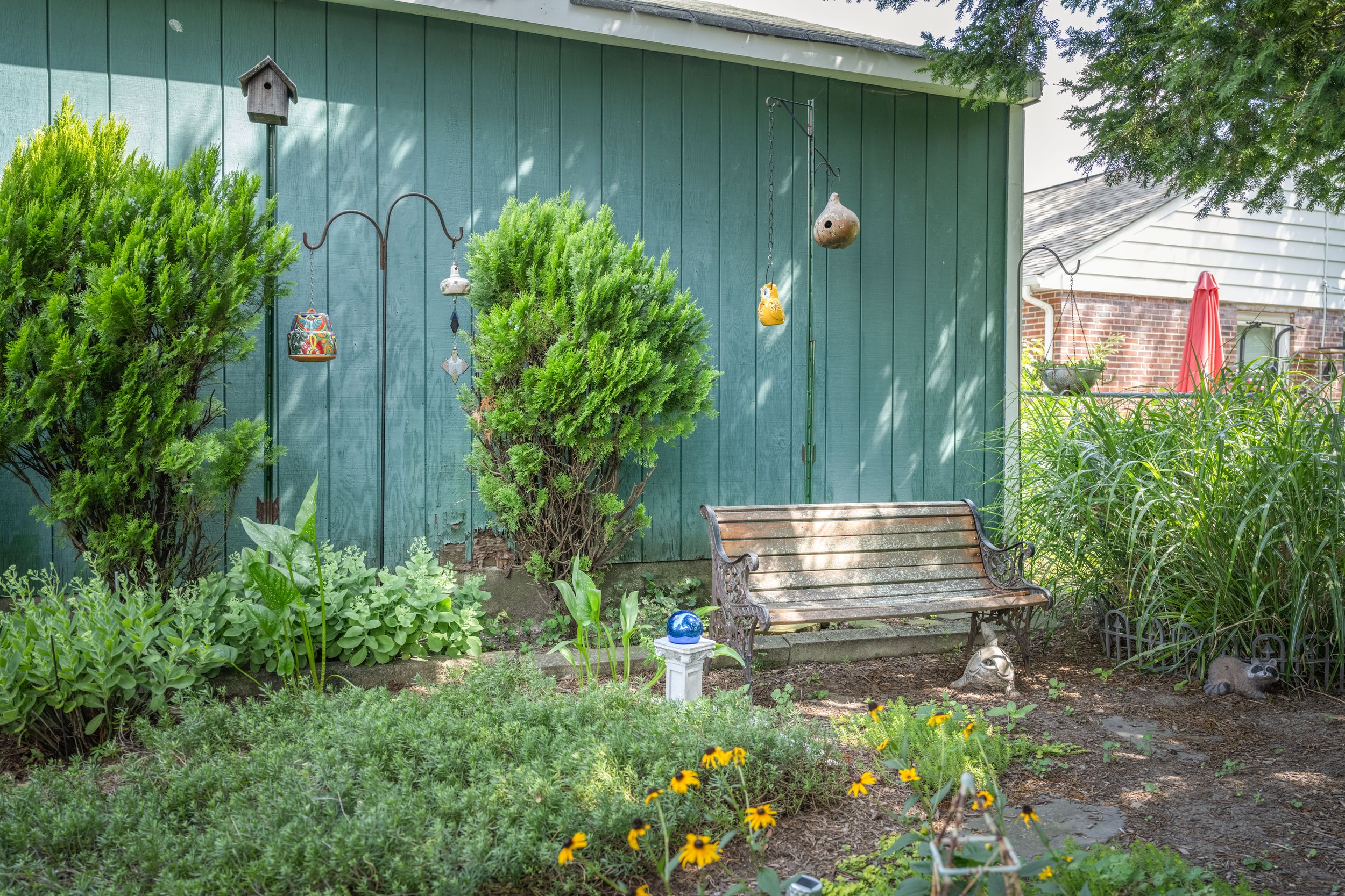A backyard garden with green trees and bushes, a green wooden wall, decorative birdhouses, hanging ornaments, a wooden bench, and yellow flowers, with a house and red umbrella in the background.