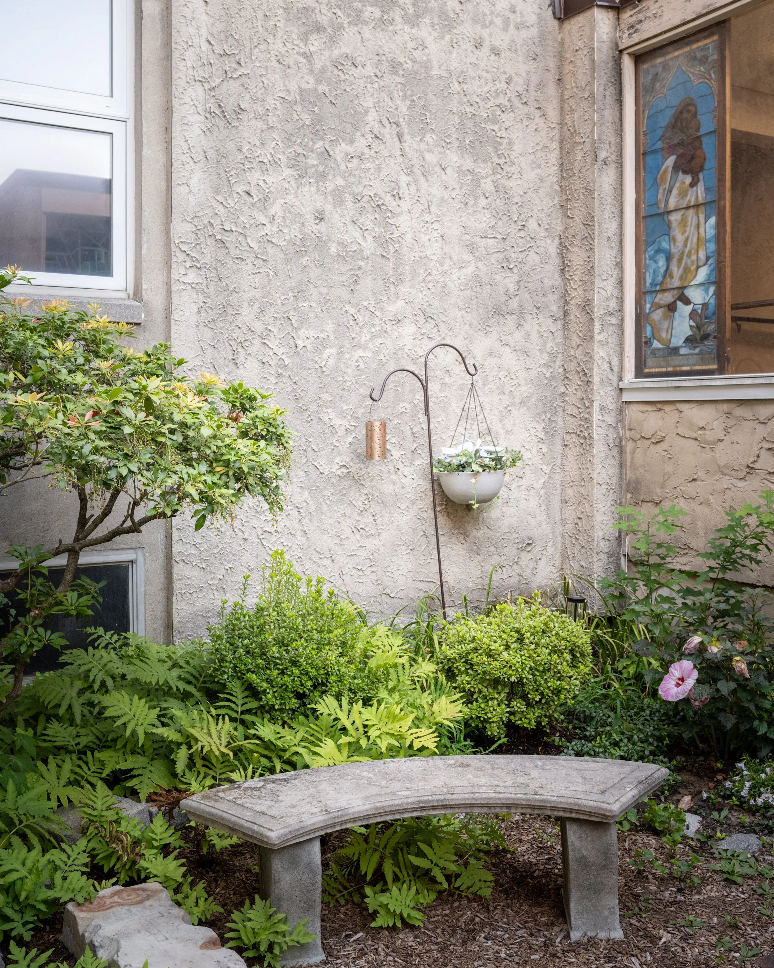 A small garden area with green plants, a stone bench, and a stucco wall with two windows. One window has stained glass depicting a woman in a yellow and blue dress holding a sword, with a blue background, and the other is a standard window. There is 