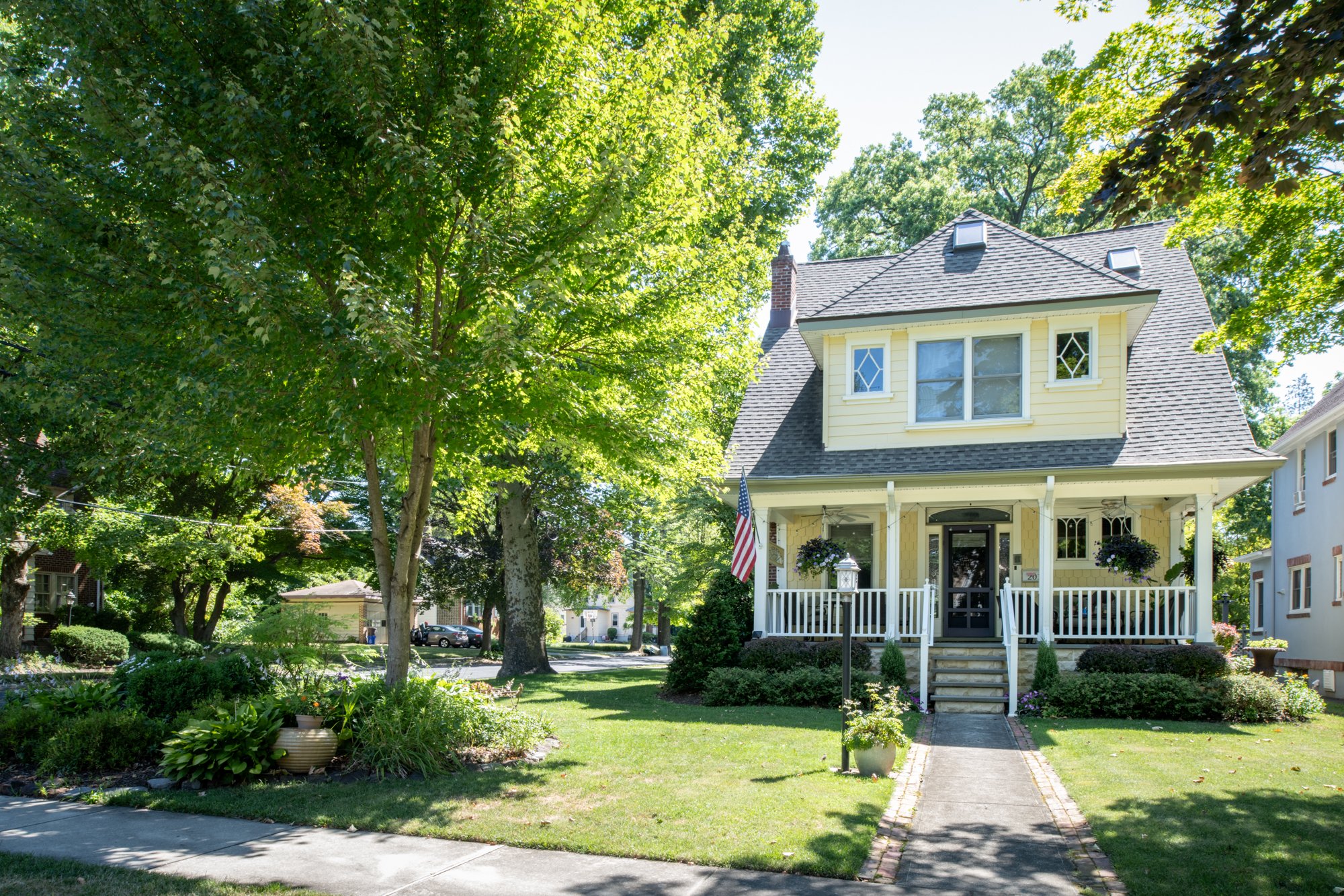 A yellow house with a porch, white railing, and potted plants, surrounded by green trees and a well-maintained lawn on a sunny day.