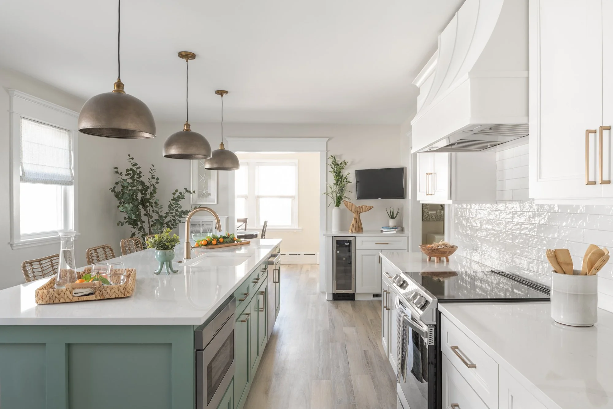 Bright modern kitchen with white cabinets, stainless steel appliances, and a large island with a white countertop. Decor includes plants, a television, and pendant lights hanging from the ceiling.