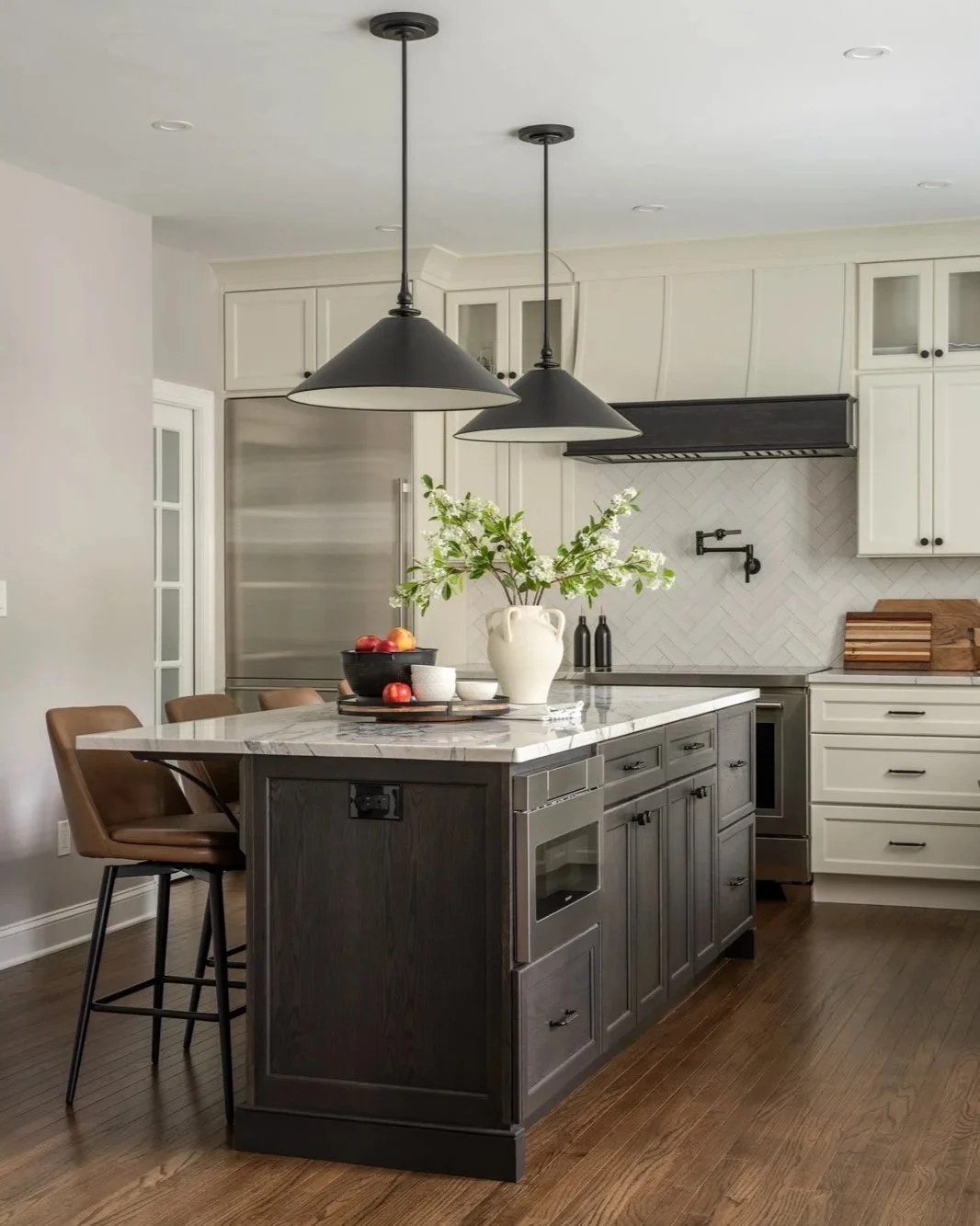 Modern kitchen with white and dark gray cabinets, a marble island with dark gray base, black pendant lights, wooden flooring, stainless steel appliances, and decorative items including a white vase with flowers and a bowl of apples.