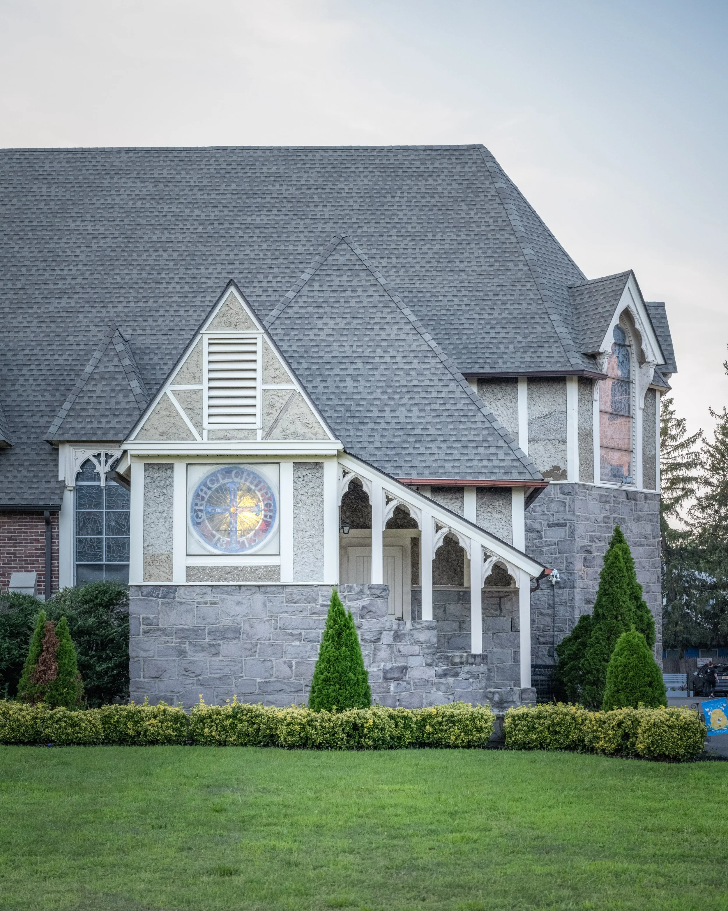 A church building with a steep, shingled roof, stained glass windows, and a front porch, surrounded by manicured bushes and green lawn.