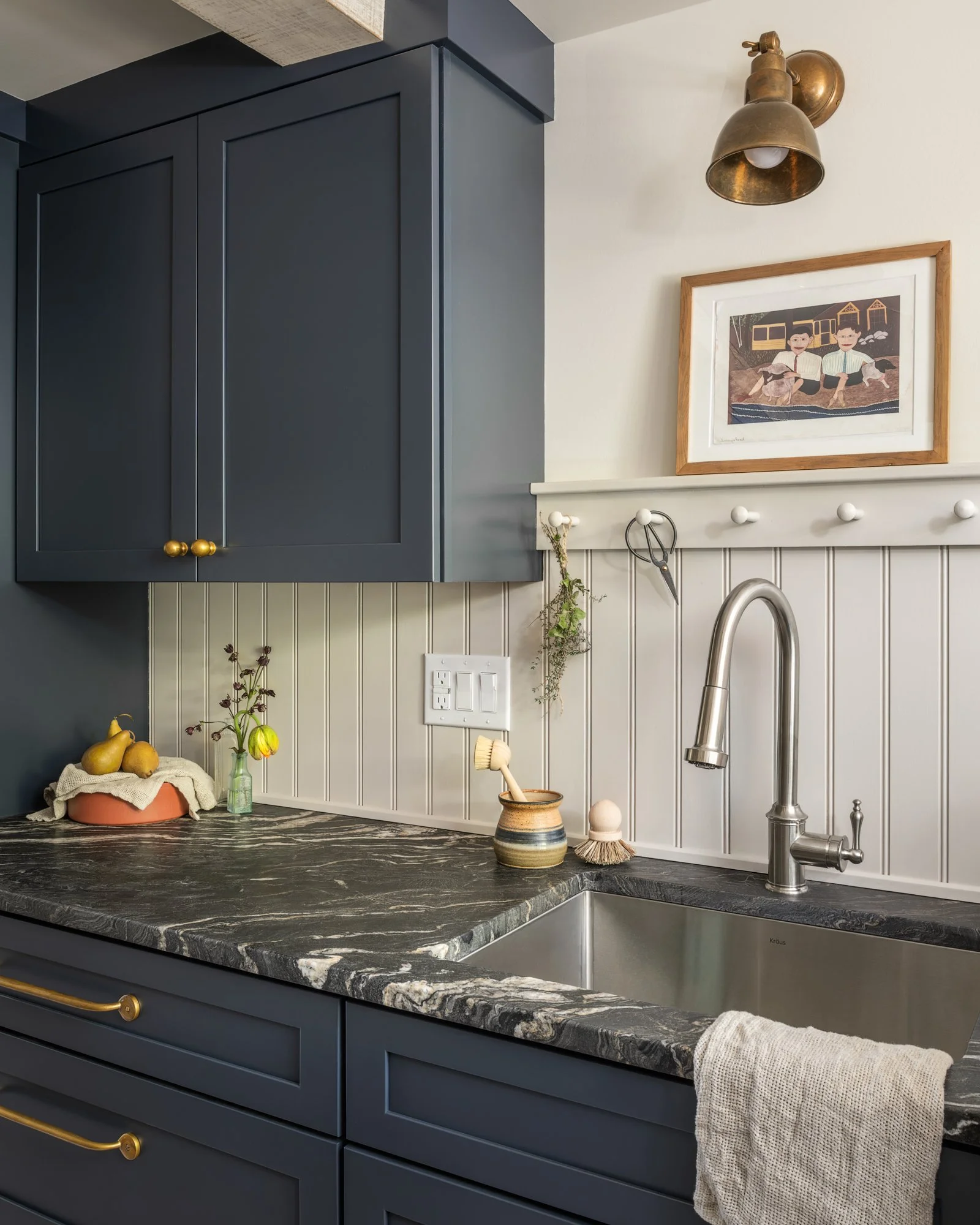 A kitchen counter with a dark marble countertop, a blue cabinet with gold handles, a stainless steel sink, a brass wall sconce light, a picture frame with a drawing of two children, and various small plants and kitchen utensils.