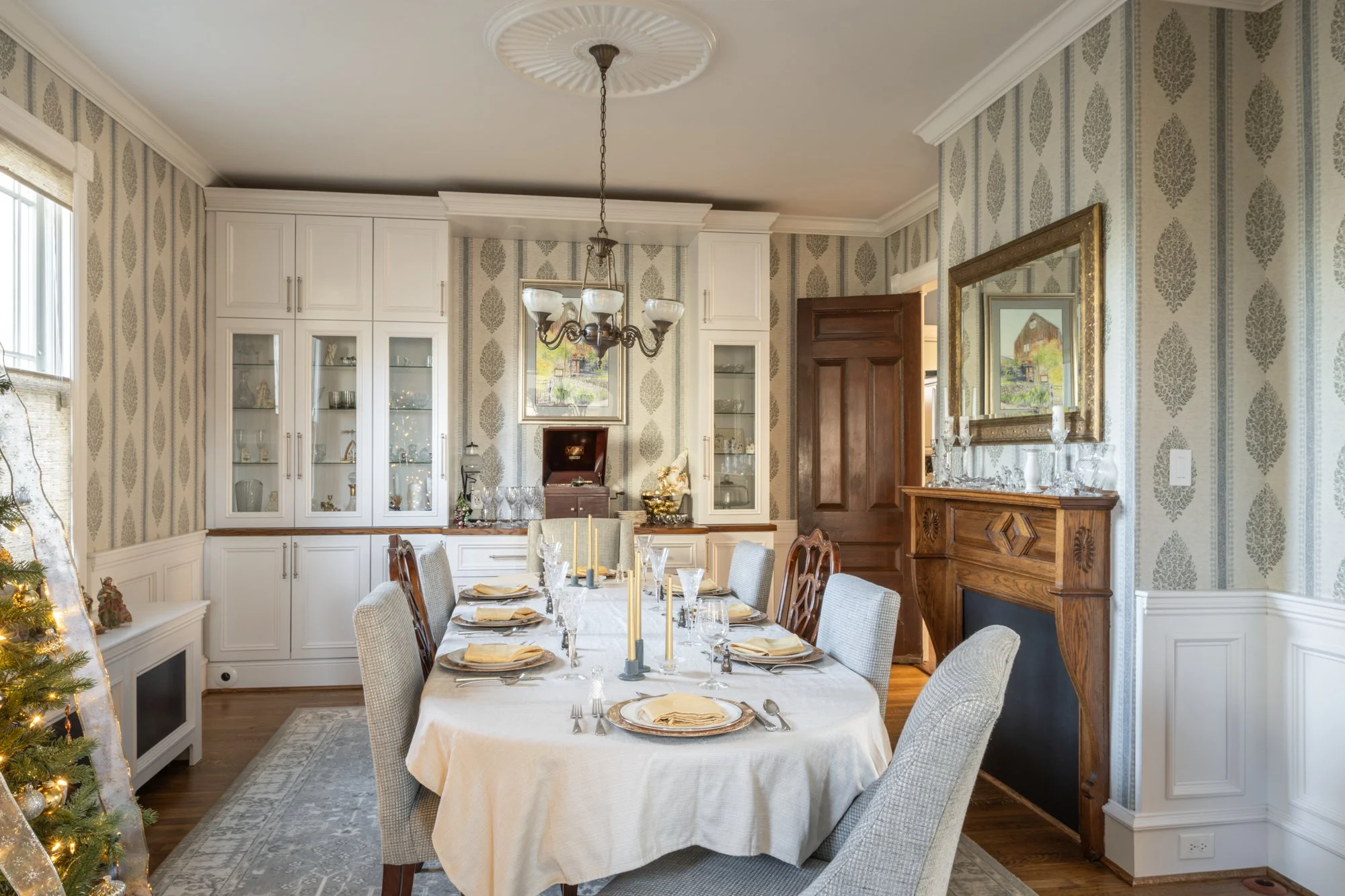 A dining room decorated for a festive occasion with a long table set with plates, gold chargers, napkins, glassware, and candles. The room features patterned wallpaper, white built-in cabinets with glass doors, a large framed mirror, and a wood firep