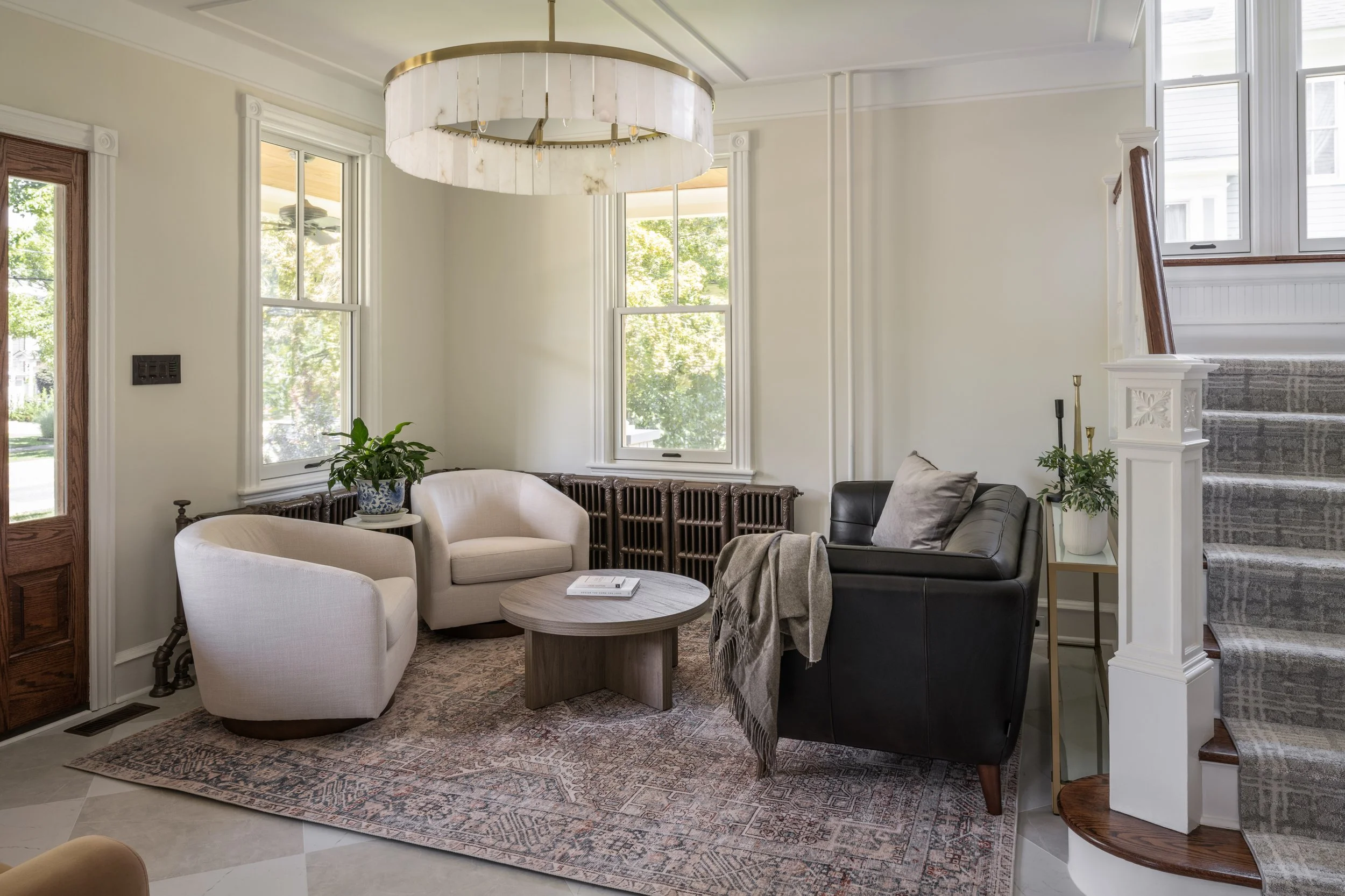 A cozy living room with natural light from large windows, featuring a white armchair, a dark leather sofa, a round wooden coffee table, and potted plants. There's a patterned area rug and a modern chandelier hanging from the ceiling, with stairs and 