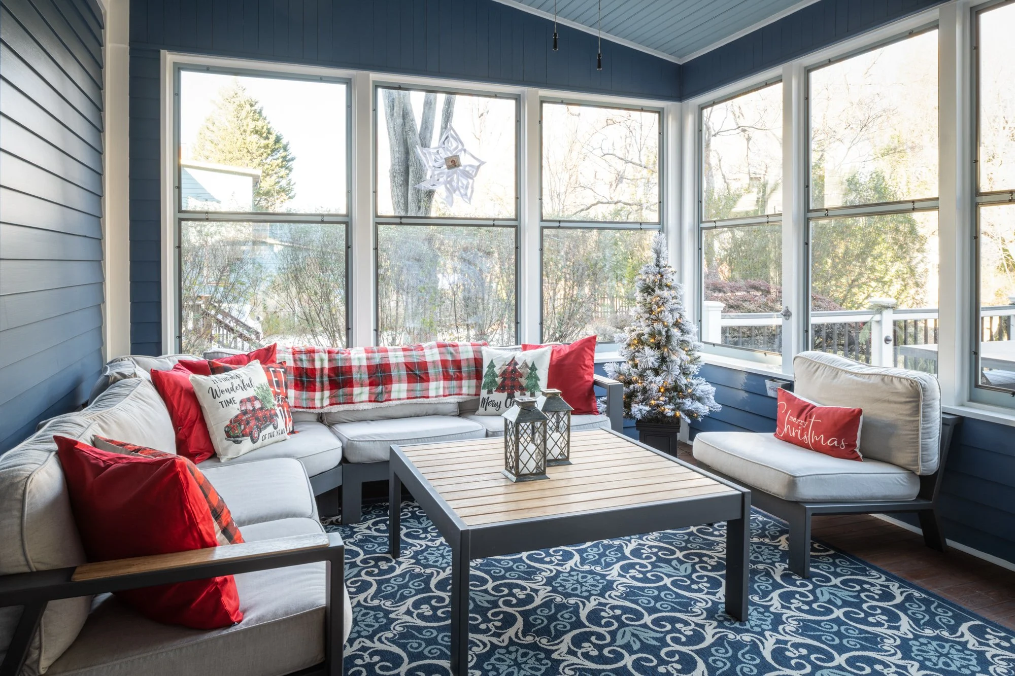 Sunroom decorated for Christmas with a small white artificial Christmas tree with lights, red and plaid throw pillows on white patio furniture, and holiday pillows with festive messages. Large windows reveal trees and a backyard outside.