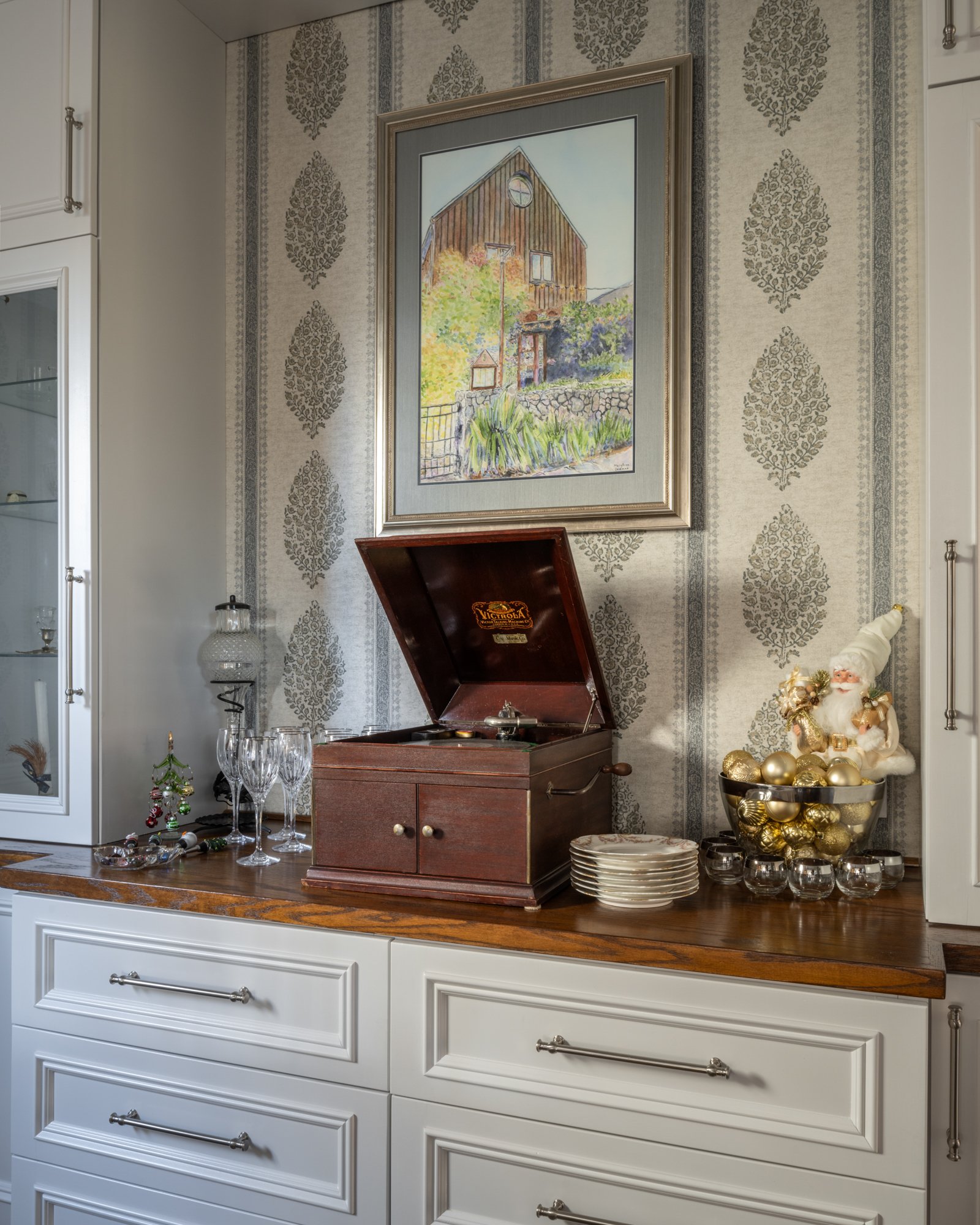 A living room sideboard with various glasses, plates, and holiday decorations. A vintage record player is on top, and a Santa Claus figurine with gold ornaments is to the right. A framed picture of a barn hangs on a decorated wallpapered wall.