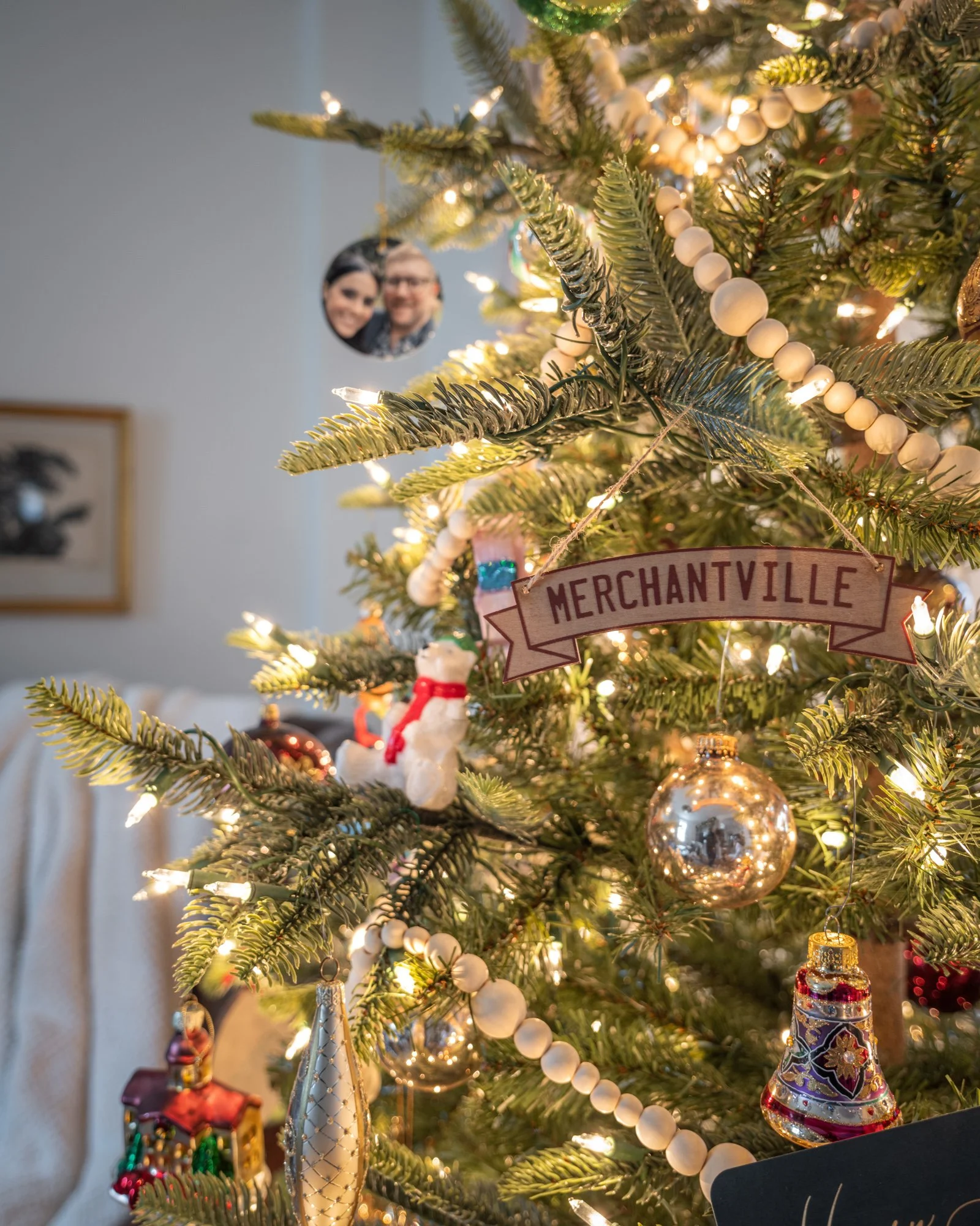 A decorated Christmas tree with various ornaments including a 'Merchantville' sign, a snowman, a bell, a colorful cone-shaped ornament, a Santa, and a small colorful gift box, illuminated with white lights.