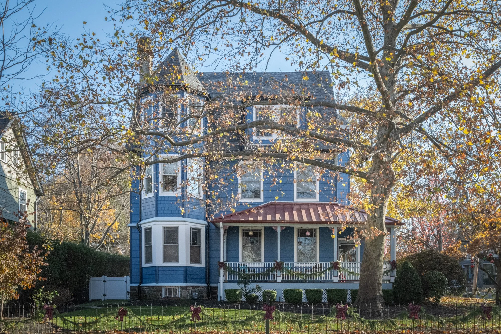 A large blue Victorian-style house with multiple gables and a bay window, surrounded by trees with autumn leaves, and a festive decorated front yard fence.