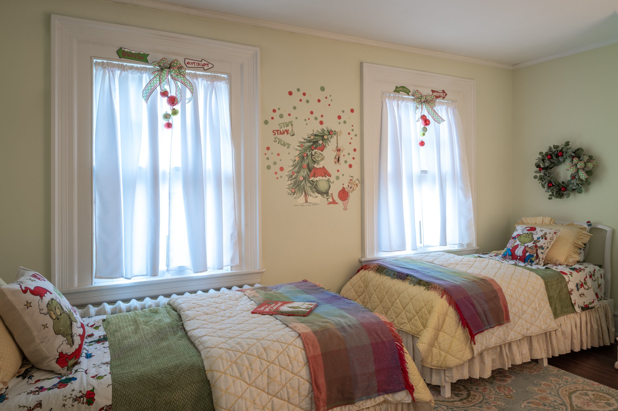 A cozy bedroom decorated for Christmas with two twin beds, each with holiday-themed bedding and pillows featuring reindeer and Santa. The wall between the beds has whimsical holiday artwork, and there is a festive wreath on the wall. Two windows with