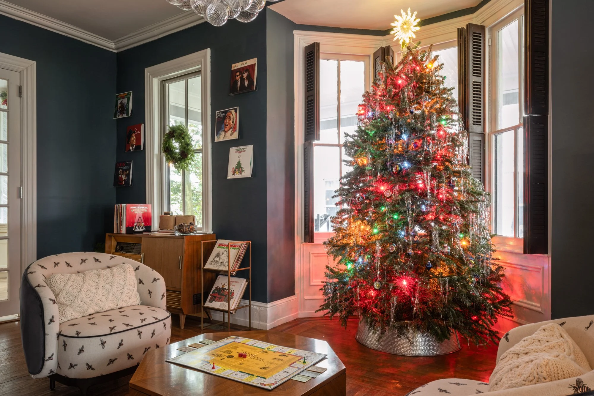 A decorated Christmas tree in a living room with windows and holiday cards on the wall.