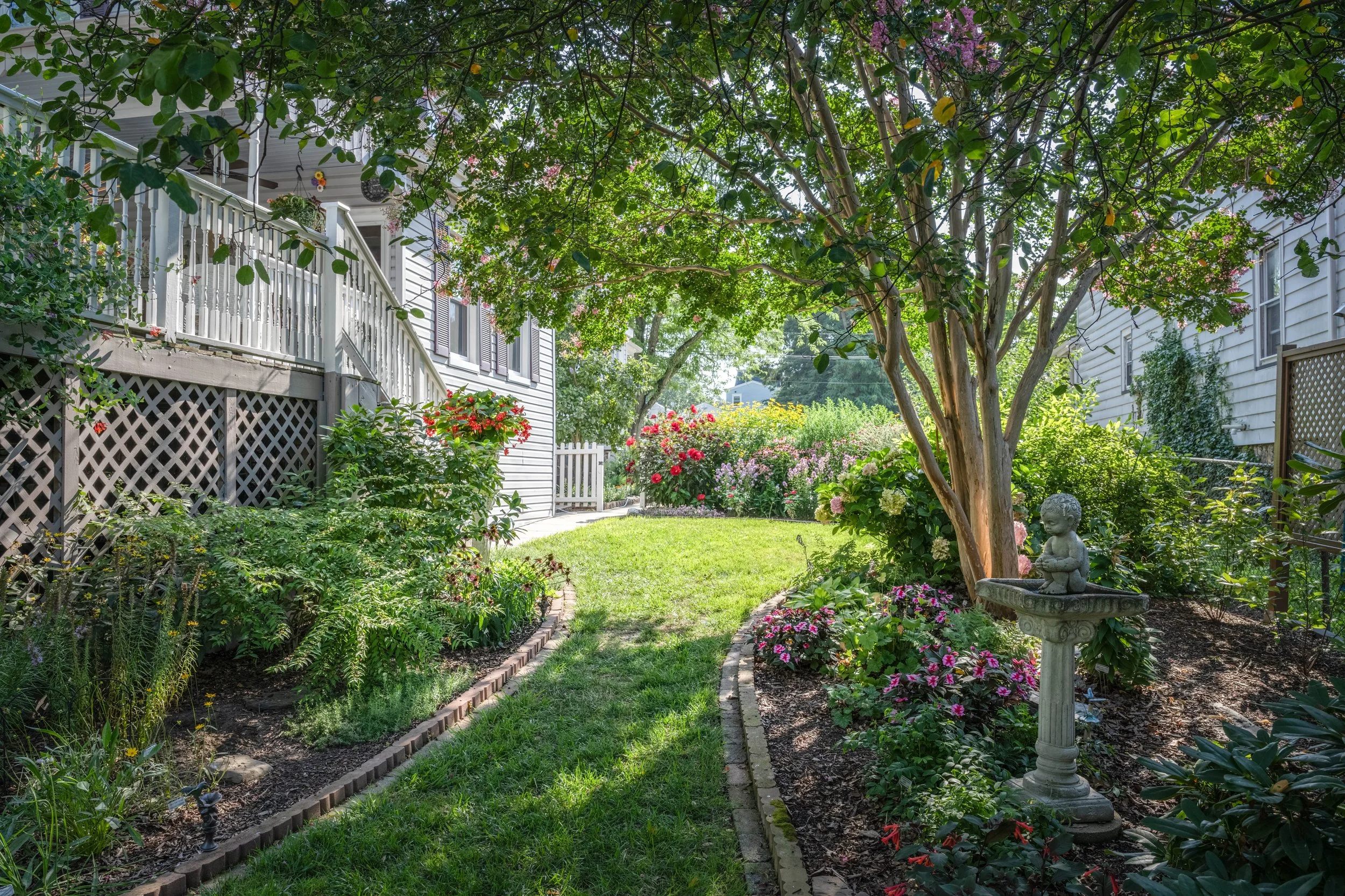 A lush garden with a brick pathway, surrounded by colorful flowers and greenery. There is a white house with a balcony and a white picket fence in the background, shaded by large trees. A stone statue of a small girl sits in the garden.