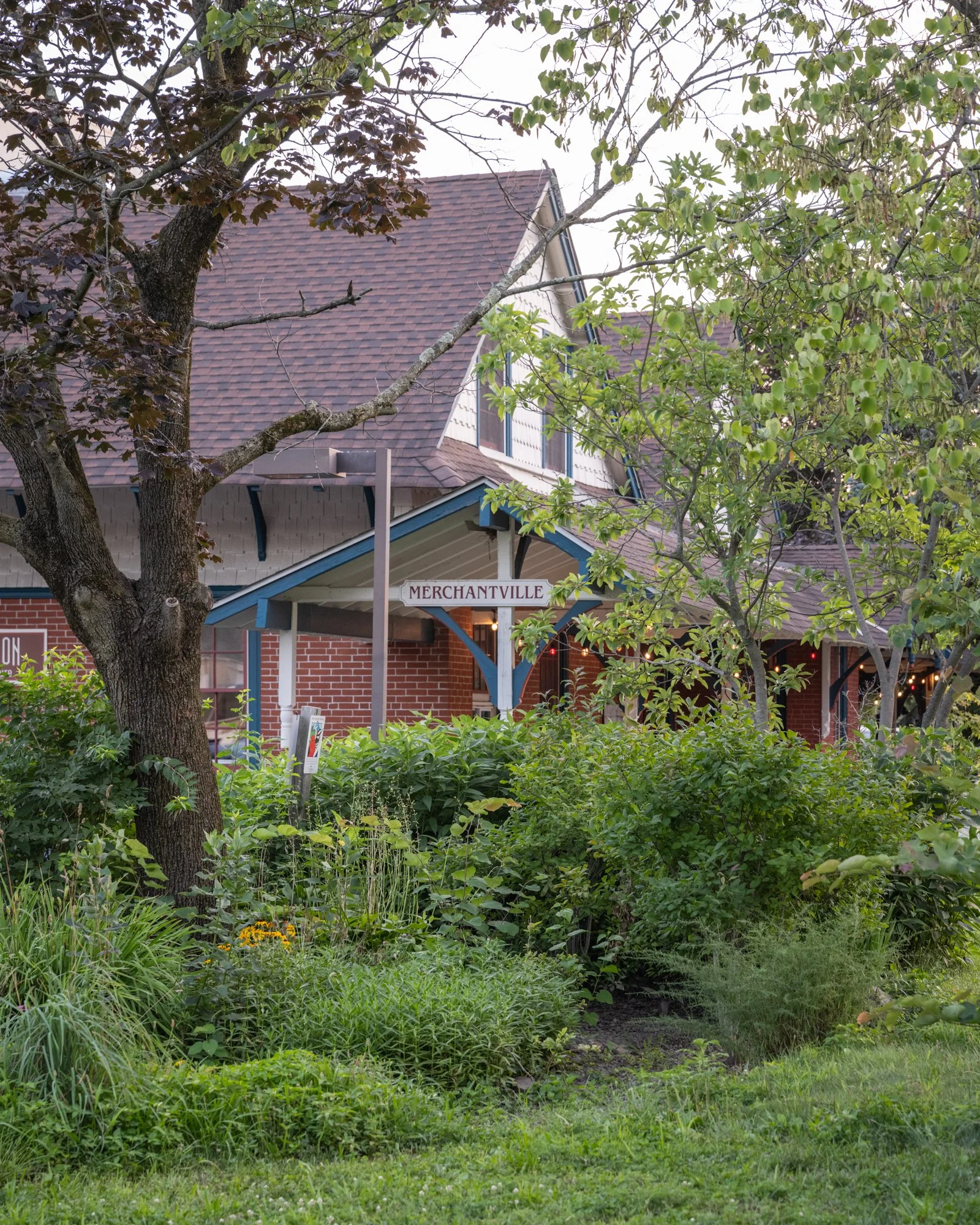 A building with a gabled roof and a sign that says "Merchantville," partially obscured by trees and lush greenery in a garden setting.