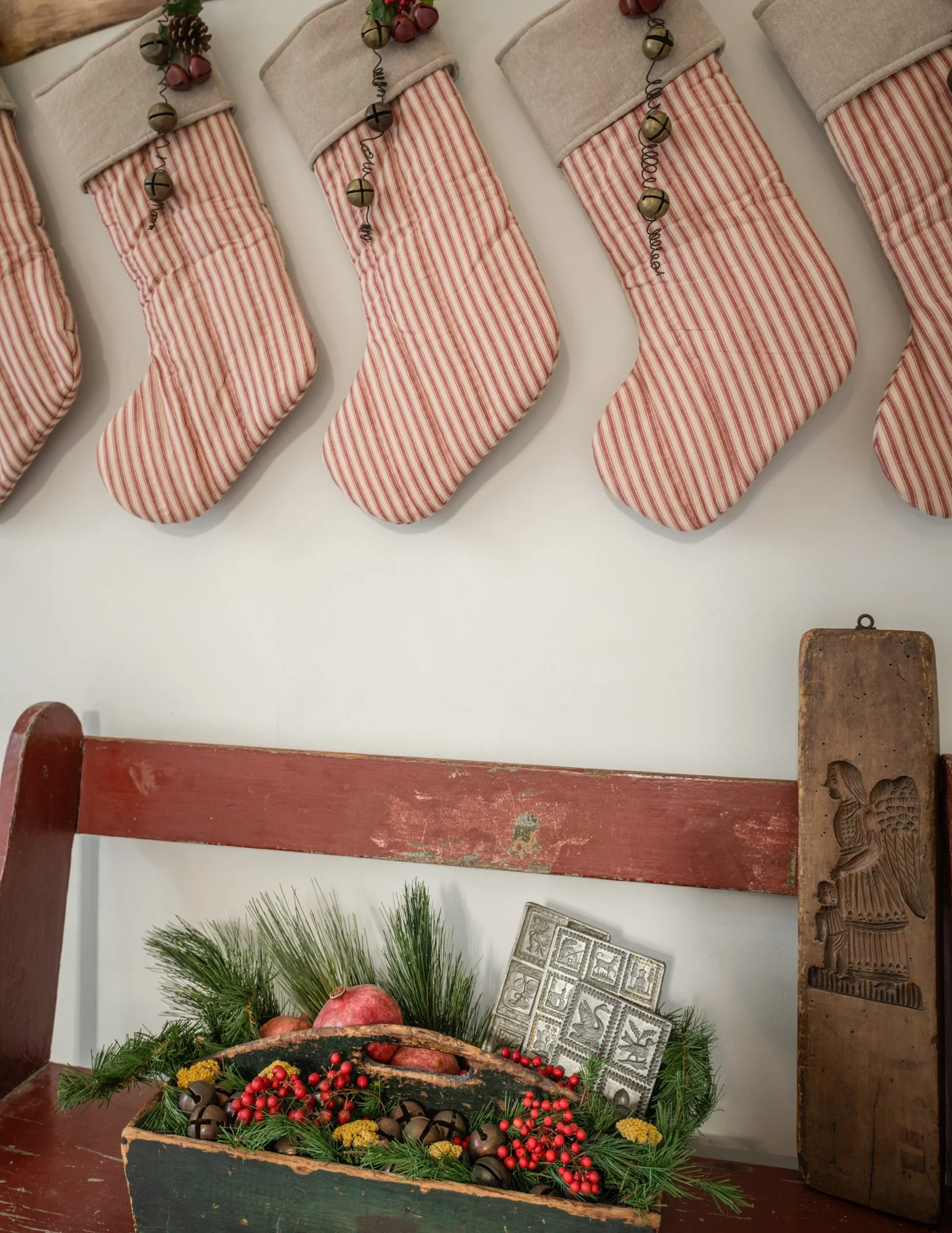 Holiday display featuring red and white striped Christmas stockings with beige cuffs hanging on a wall, a vintage red wooden bench with pine branches, red berries, yellow flowers, and a rustic wooden box filled with pine branches and ornaments, along