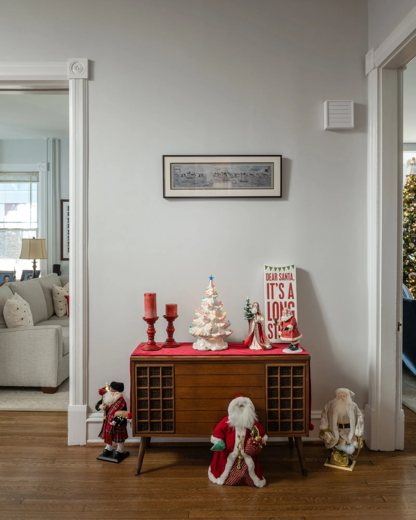 Living room with a side table displaying Christmas decorations, including a miniature Christmas tree and Santa figurines, with holiday-themed decor on the wall and ornaments.