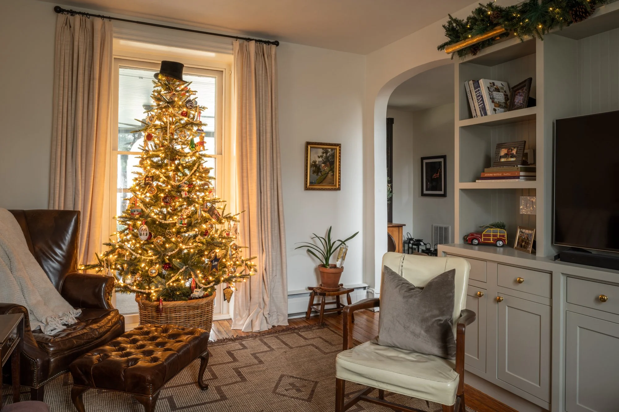Living room with a decorated and lit Christmas tree near a large window with beige curtains, a potted plant, a framed picture on the wall, and a cabinet with books and decorative items.