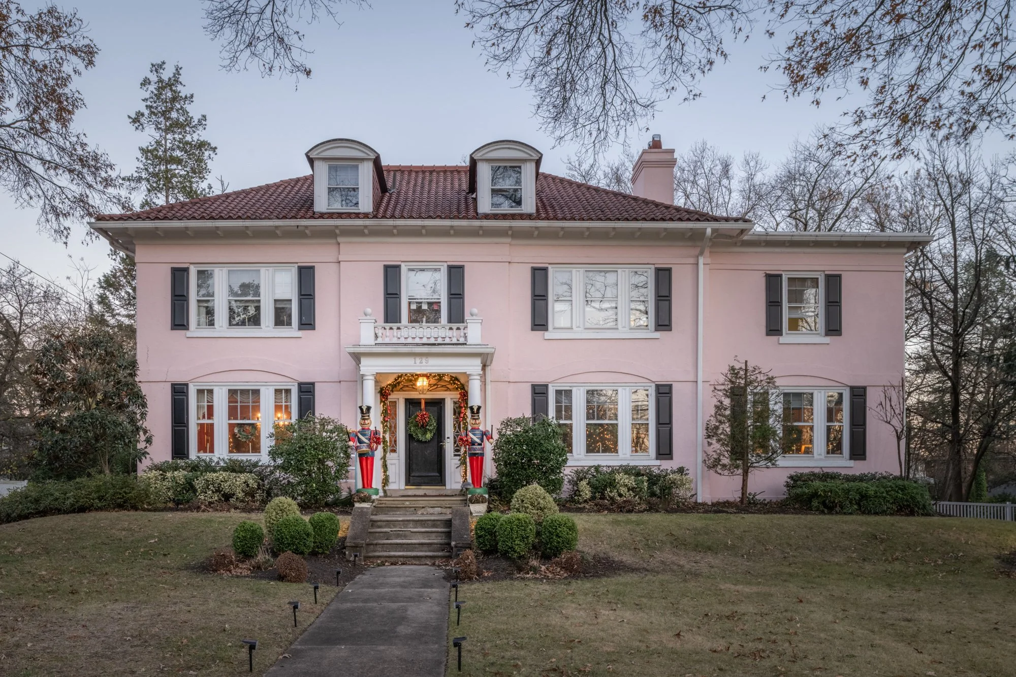 A large, pink, three-story house decorated for Christmas with a wreath on the front door, garlands, and toy soldiers flanking the steps. The house has white trim, black shutters, and a red-tiled roof. The front yard has a walkway, bushes, and trees w