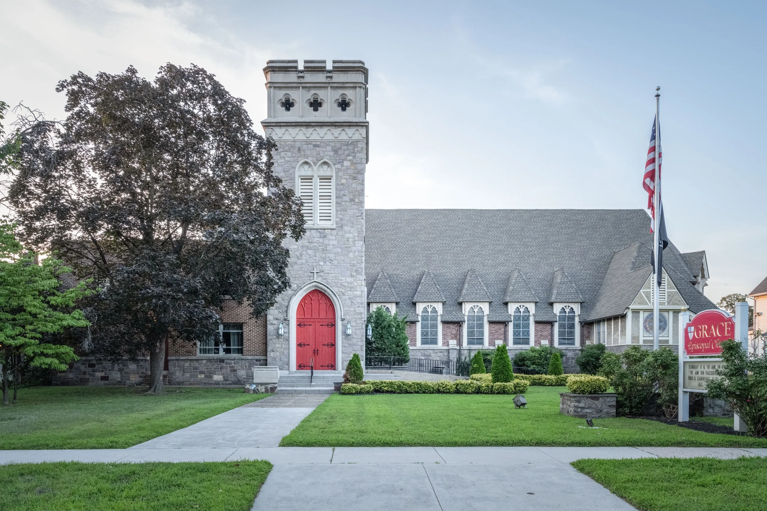 A church with gray stone and brick walls, red doors, and a tall tower with three gothic arches, surrounded by green grass, trees, and a sidewalk, with American flags and a church sign in front.