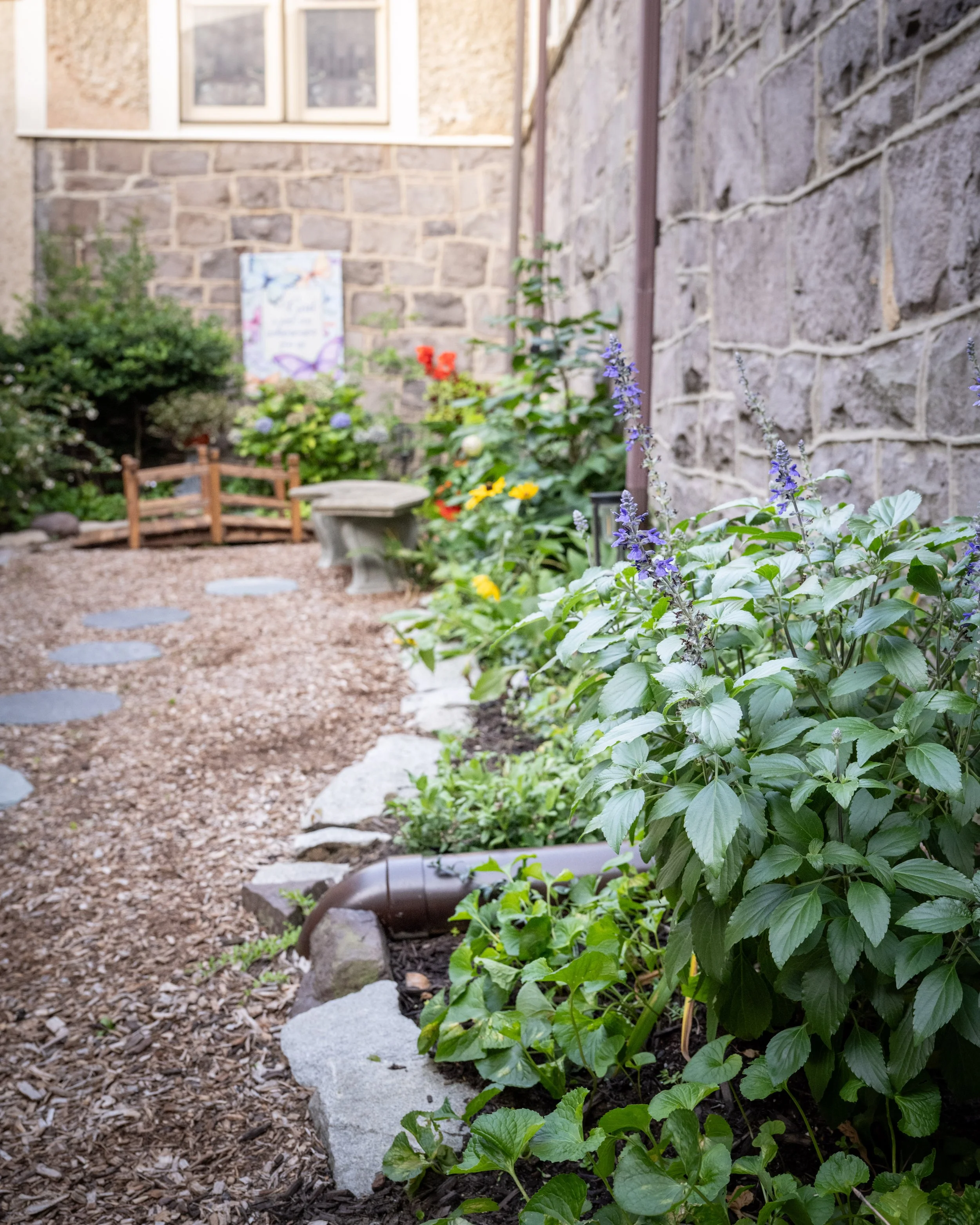 A garden with flower beds, bushes, and plants, along with a stone and gravel pathway, two small stone benches, a brick wall, a window, and a poster on the wall.