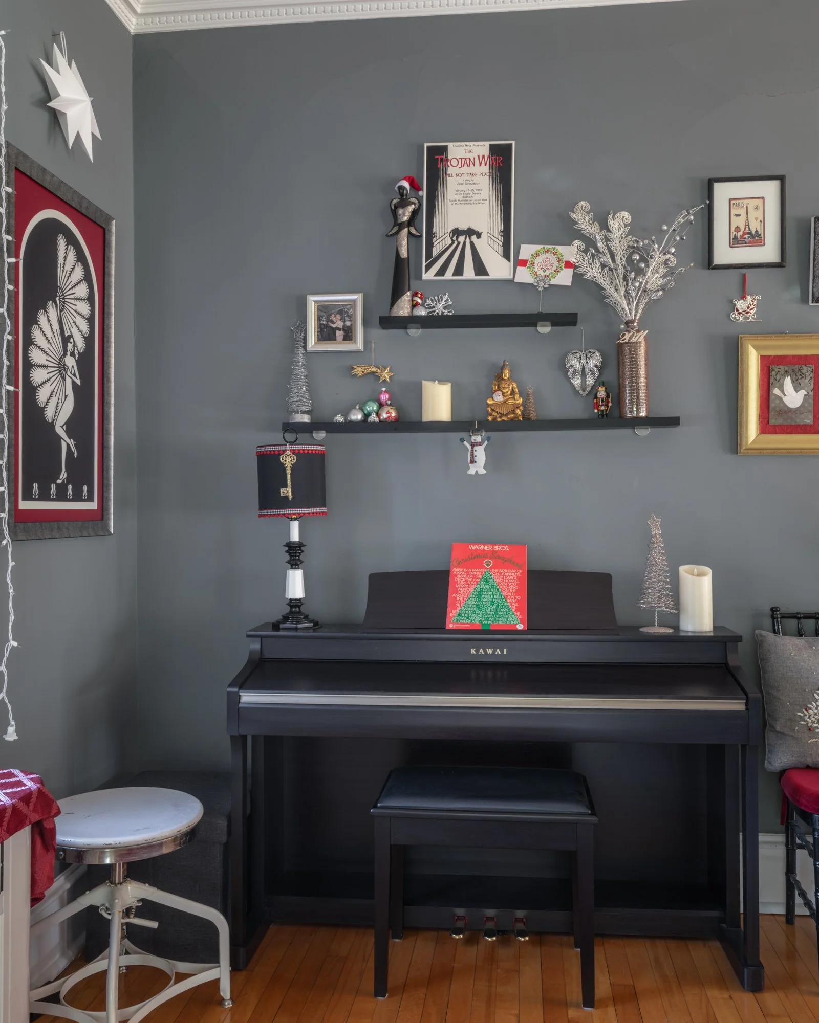 A black digital piano with a small red and green holiday card on top, placed against a gray wall decorated with various holiday ornaments and pictures, including a Buddha statue with a Santa hat, holiday-themed posters, and a white decorative branch 