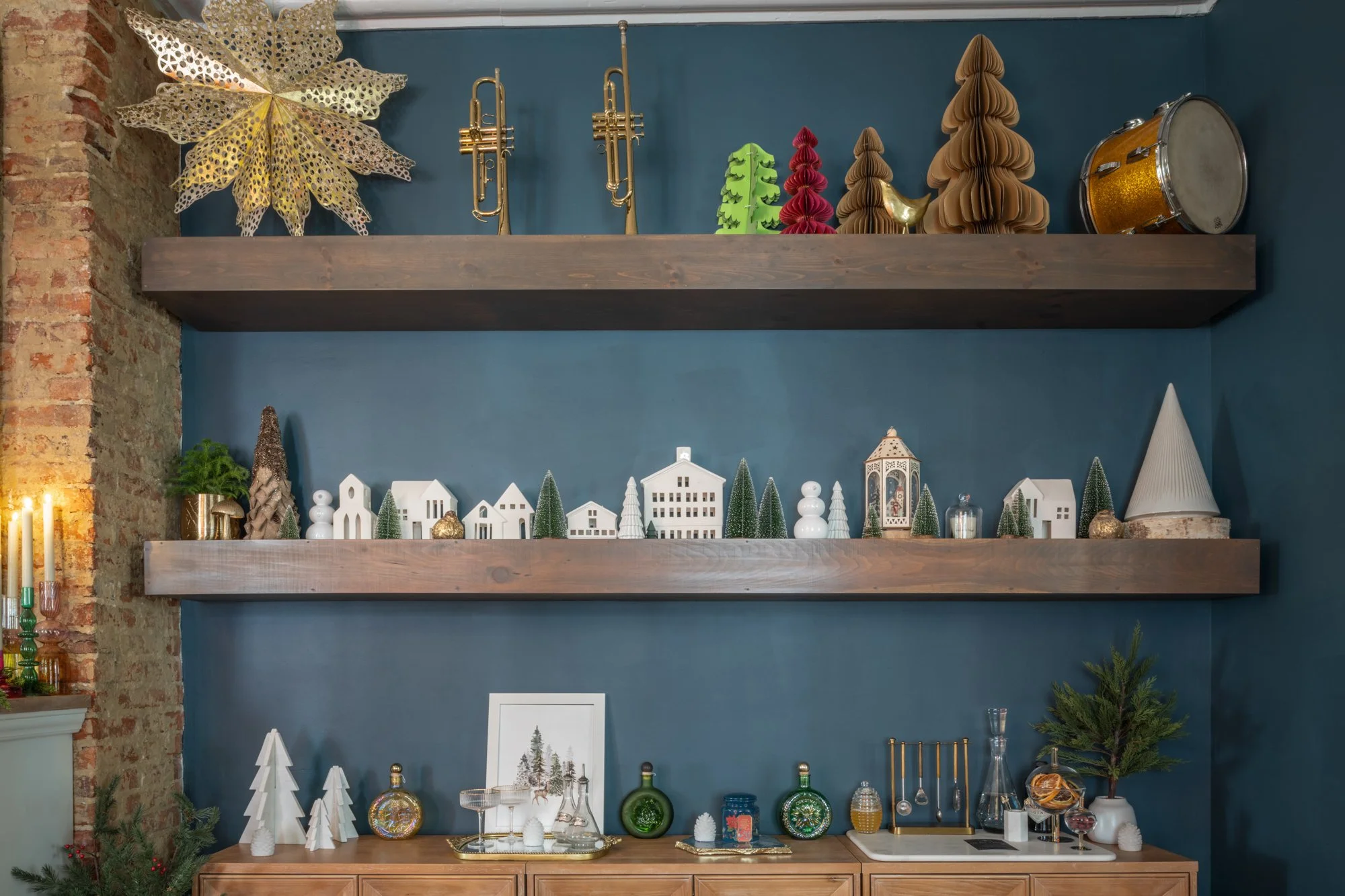 Display of holiday decorations including miniature Christmas trees, glass ornaments, and winter-themed figurines on wooden shelves and a sideboard against a teal wall.