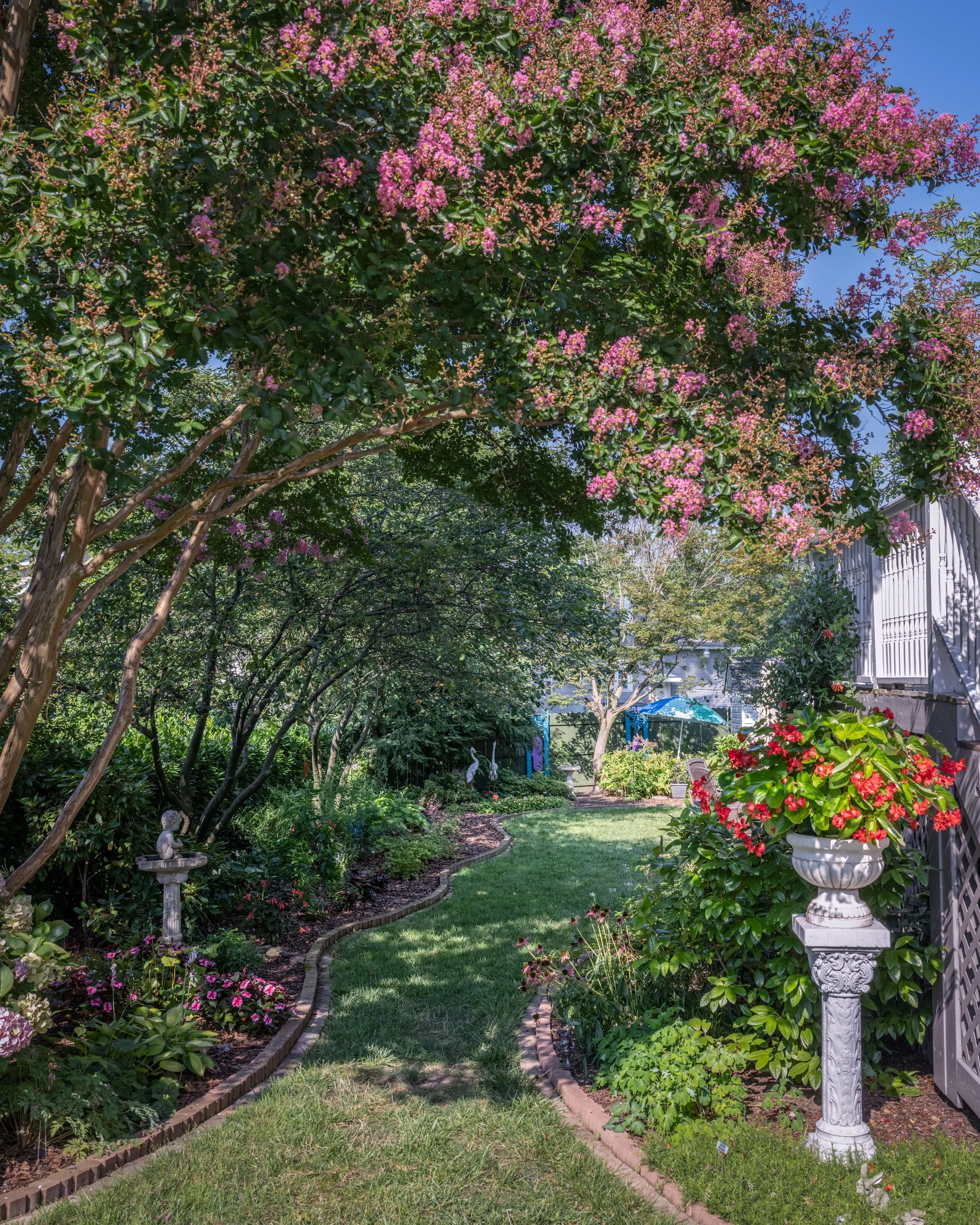 A lush garden scene with a brick-edged lawn, colorful blooming flowers, a white decorative bird bath, and a white pedestal with a large flower pot filled with vibrant red and pink flowers. A shaded pathway curves through the garden, partially visible
