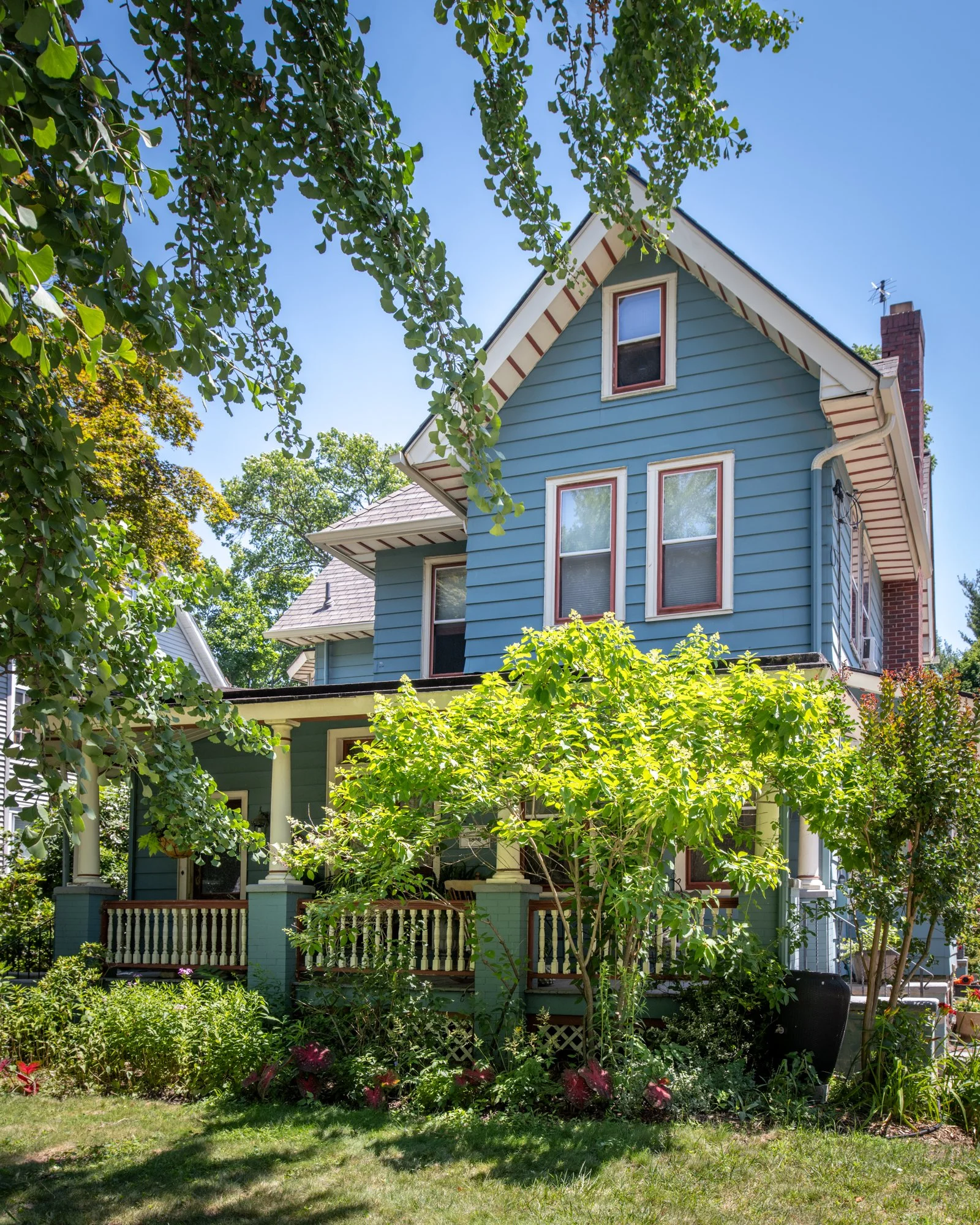 A blue two-story house with white trim, multiple windows, and a brick chimney, surrounded by green trees and shrubs under a clear blue sky.