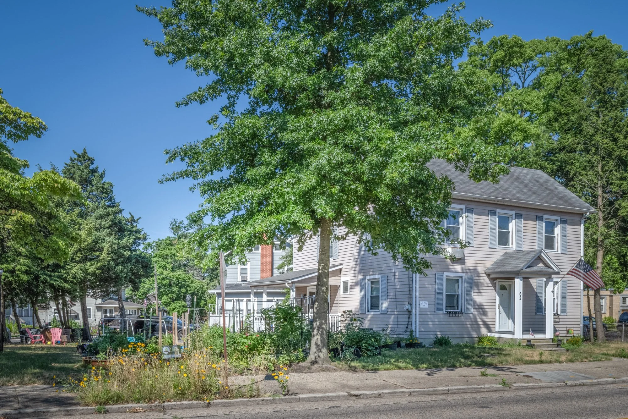 A two-story house with beige siding, gray roof, and white window shutters, surrounded by green trees and a garden with colorful chairs and flowers, under a clear blue sky.