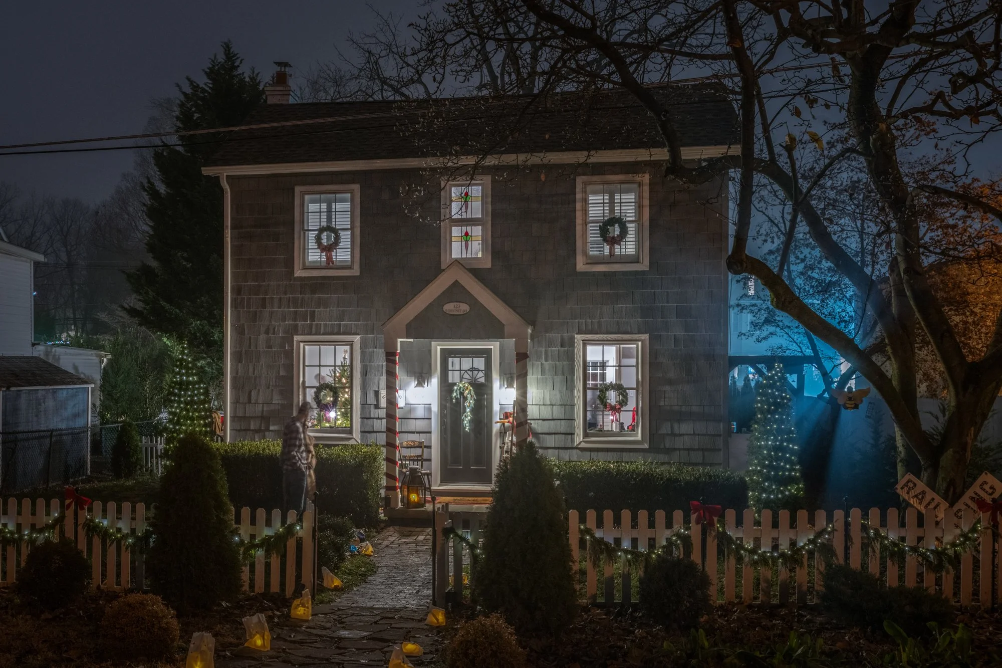 Front view of a house decorated with Christmas wreaths on the windows and Christmas lights, with a white picket fence, trees, and a game outside, at night.