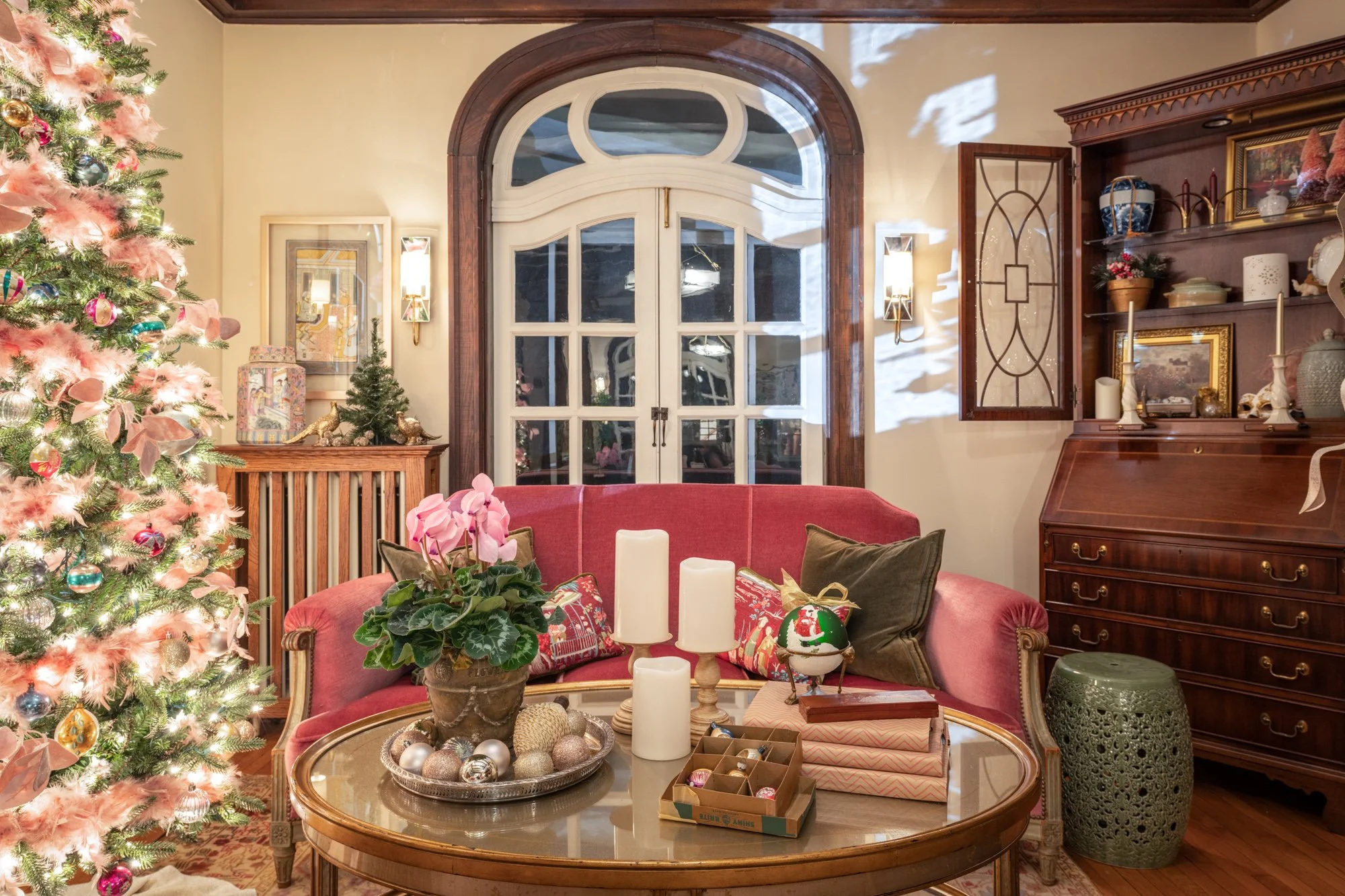 A cozy living room decorated for Christmas with a large lit Christmas tree on the left, a pink couch in the center with pillows and holiday decorations, a wooden cabinet on the right with various decorative items, and a window with rounded wood trim.
