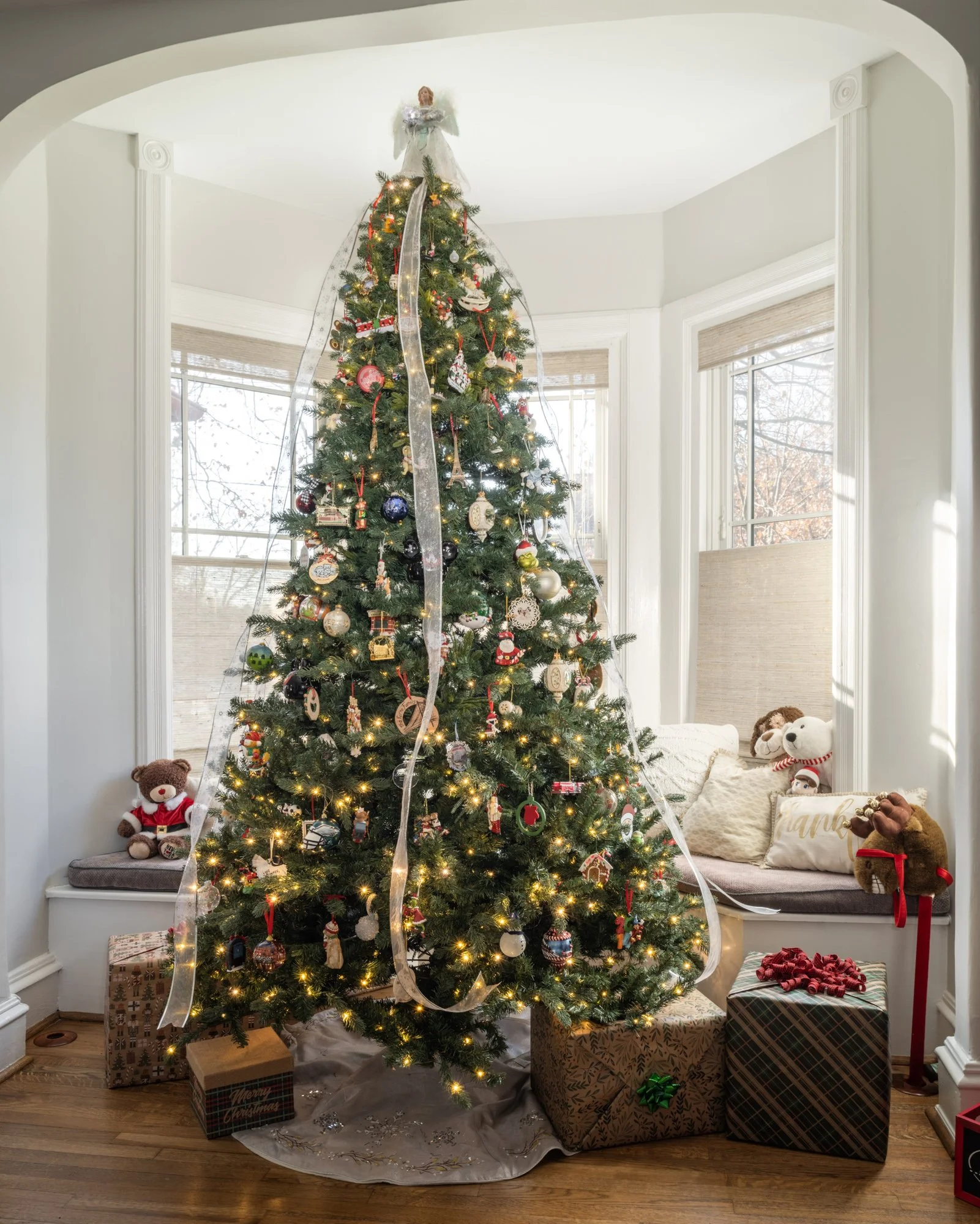 A decorated Christmas tree with ornaments, lights, and an angel on top, surrounded by wrapped presents, with a cozy seating area with plush toys and decorative pillows nearby.
