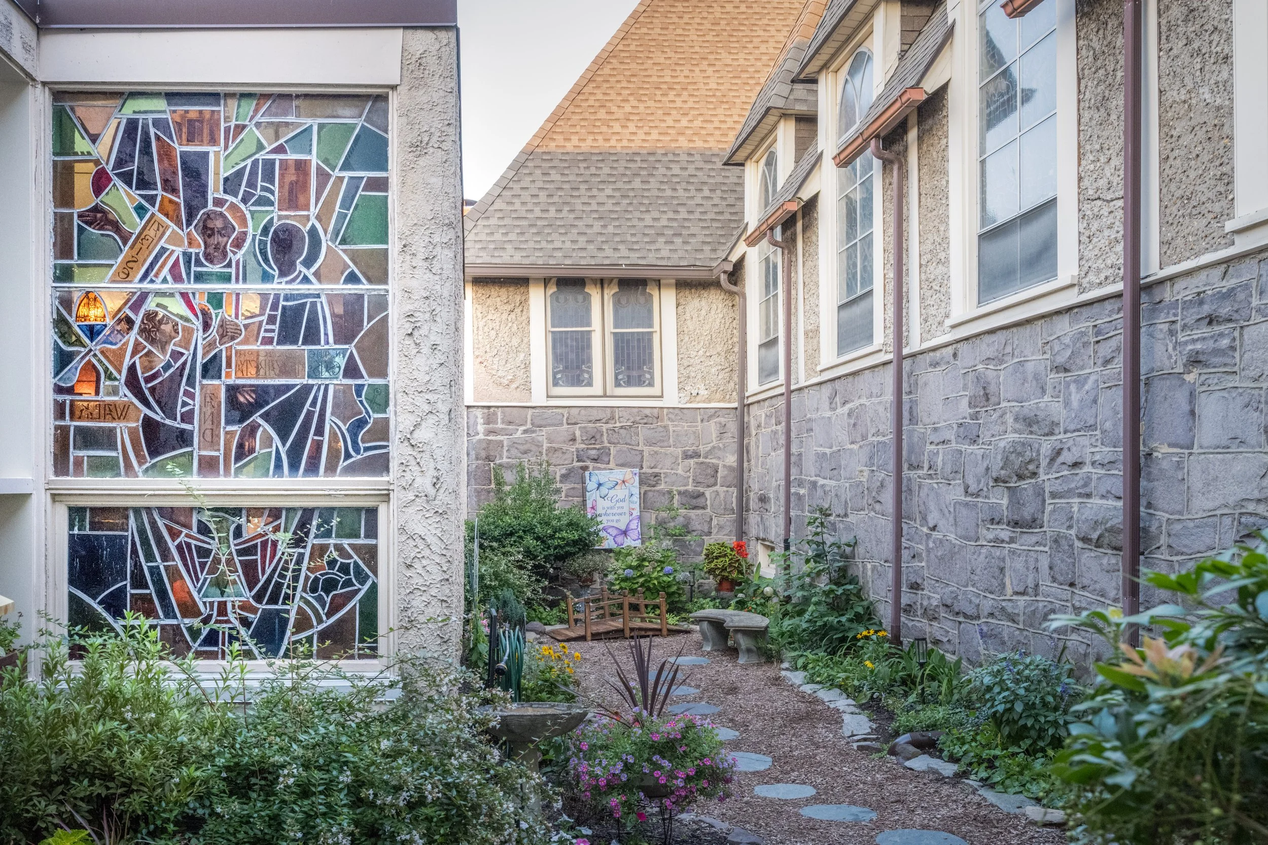 A garden with flowers and plants, a stone bench, and a small pathway under a residential house with large windows and a stained glass window on the side wall.