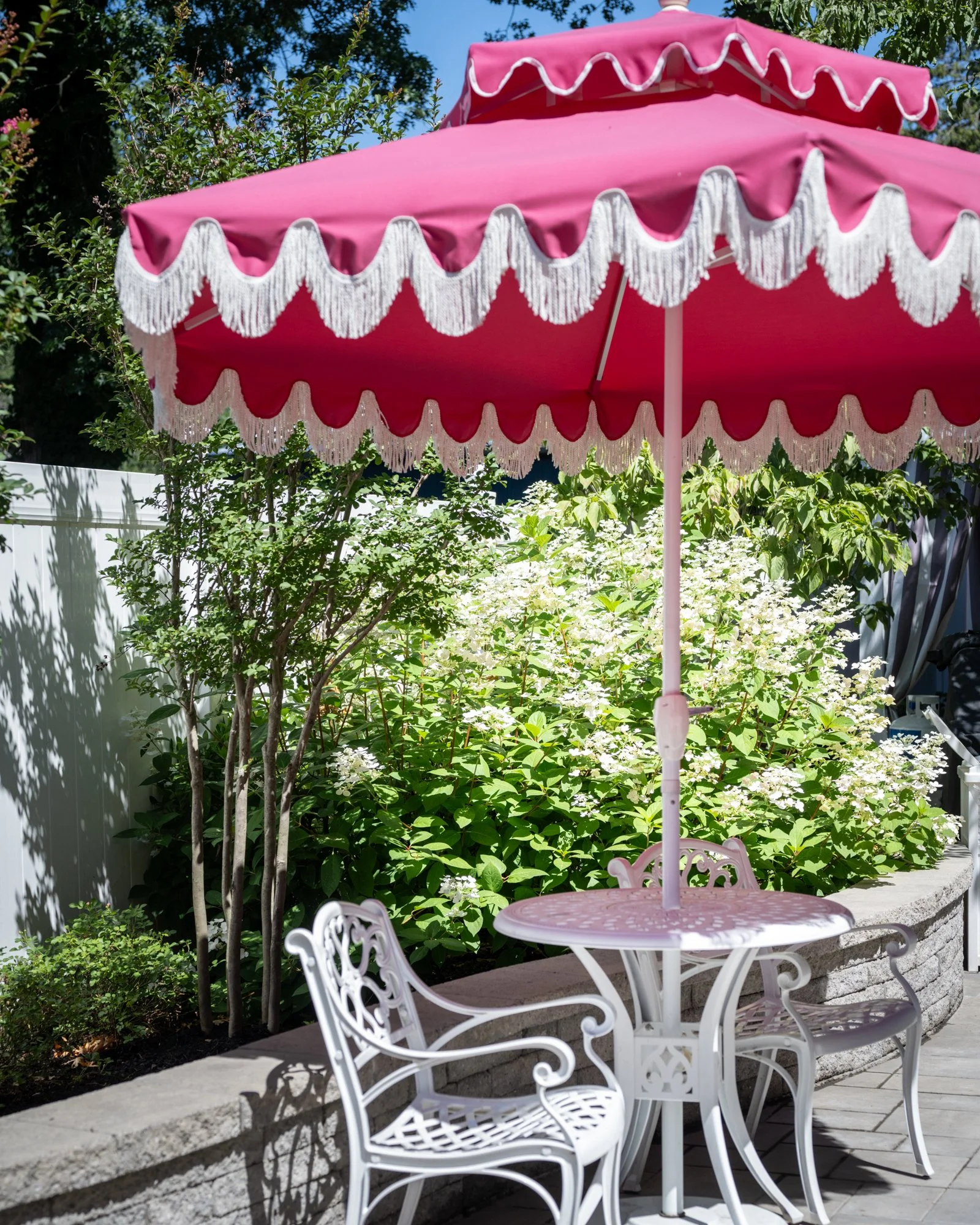Pink patio umbrella with white fringe over a white metal table and chairs on a garden patio surrounded by green shrubs and trees.