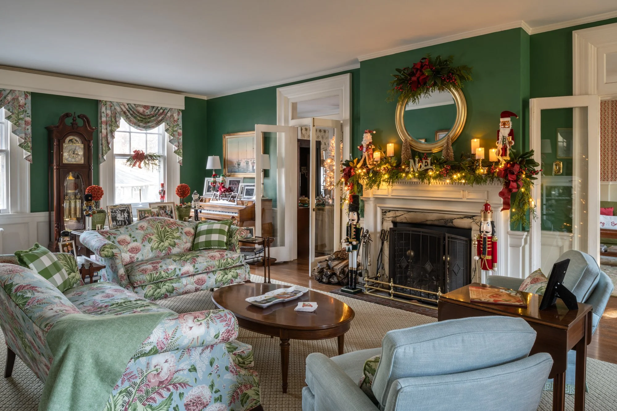 Living room decorated for Christmas with a green wall, floral sofas, and a fireplace adorned with candles, nutcracker figures, and holiday greenery. There is a large mirror above the fireplace and windows with floral curtains.
