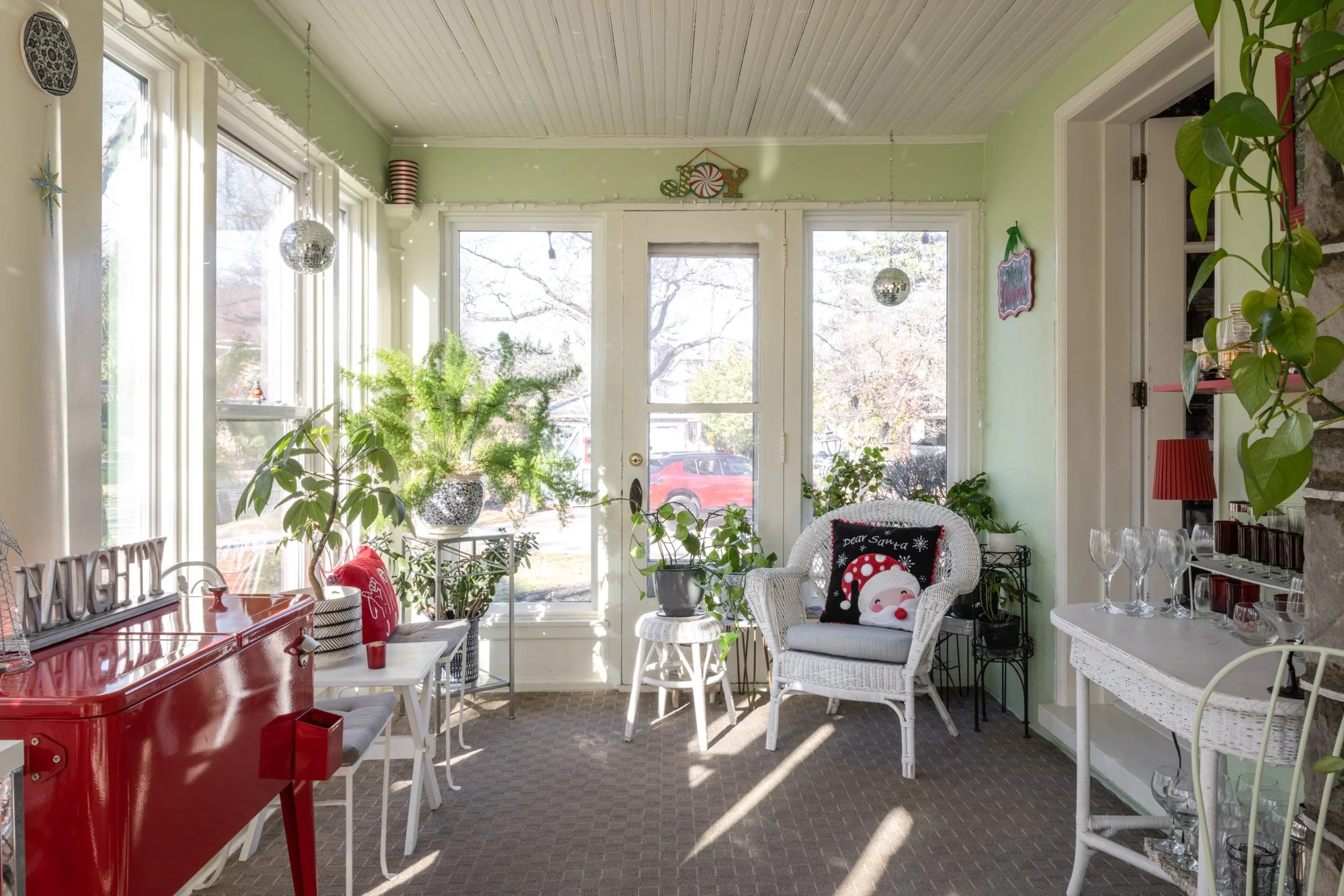 Sunlit enclosed porch with green walls, white wicker chairs, potted plants, holiday decor including Santa pillow and 'Joy' decoration, and a red vintage sewing machine on a table.