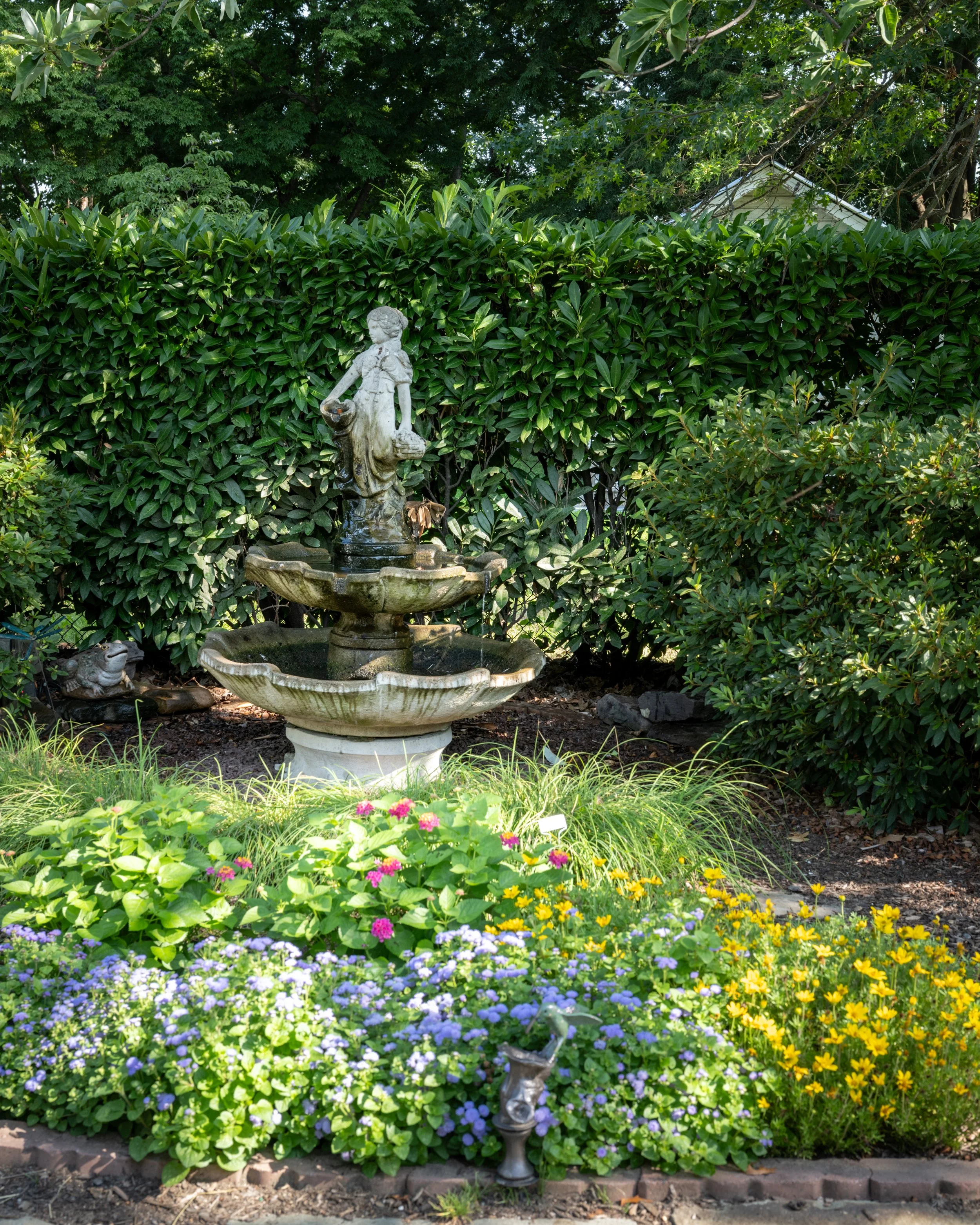 A decorative garden fountain with a statue of a girl, surrounded by lush green bushes and colorful flowers, in a sunny outdoor setting.