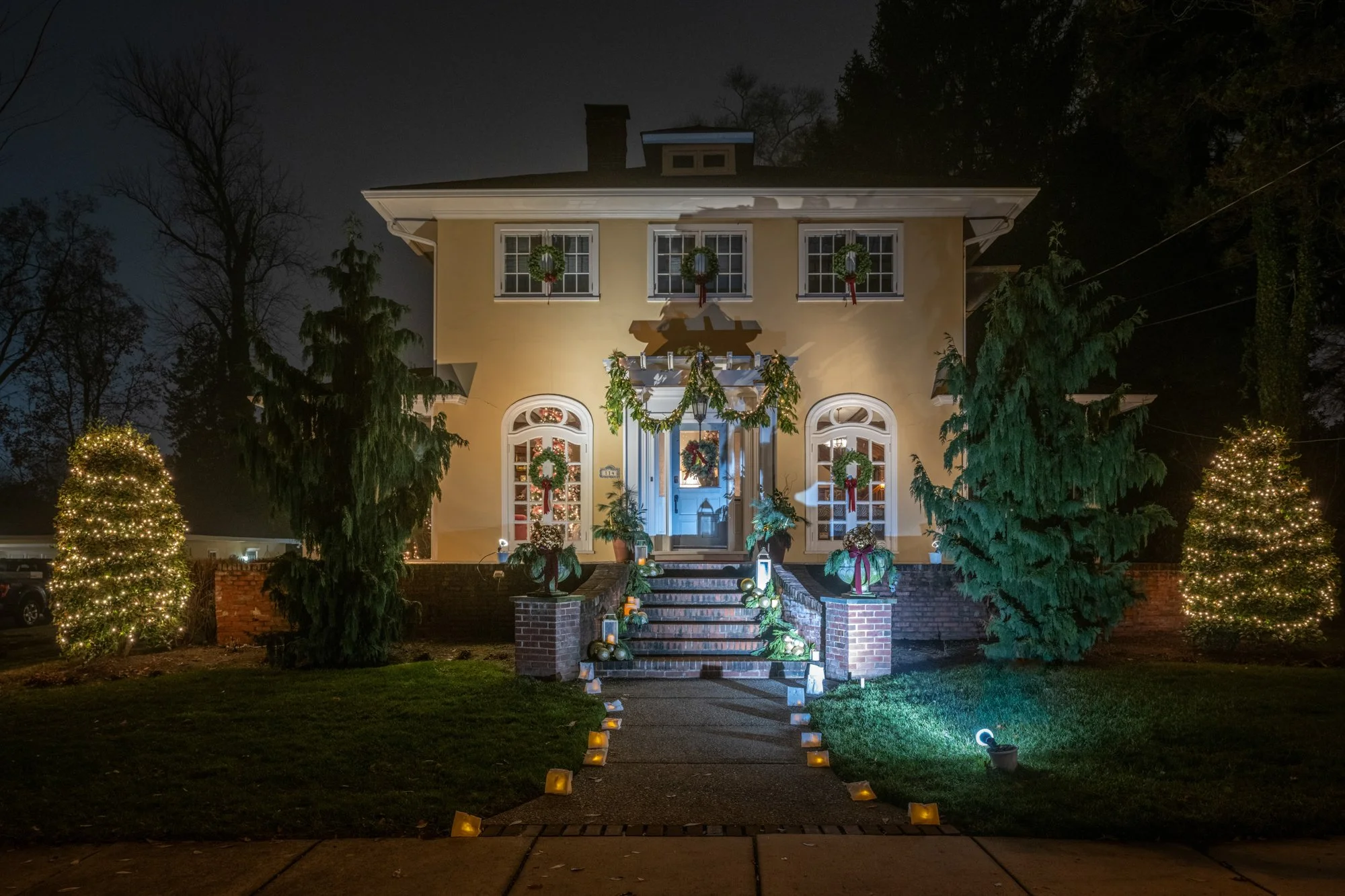 A two-story house decorated with Christmas wreaths, lights, and ornaments. The front yard has lit-up Christmas trees and a pathway with candles leading to the front steps. The house is illuminated against a dark night sky with bare trees in the backg