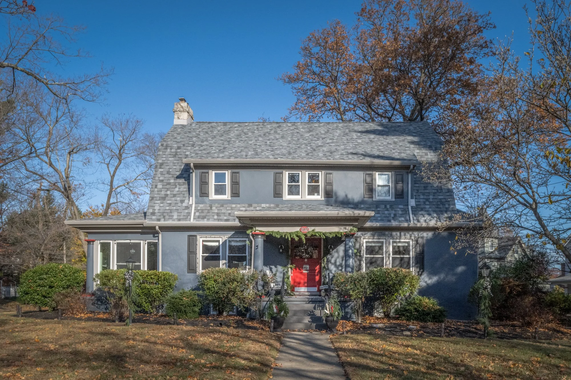 A gray house with black shutters and a red front door, decorated with greenery and holiday decorations, surrounded by trees with autumn leaves, under a clear blue sky.