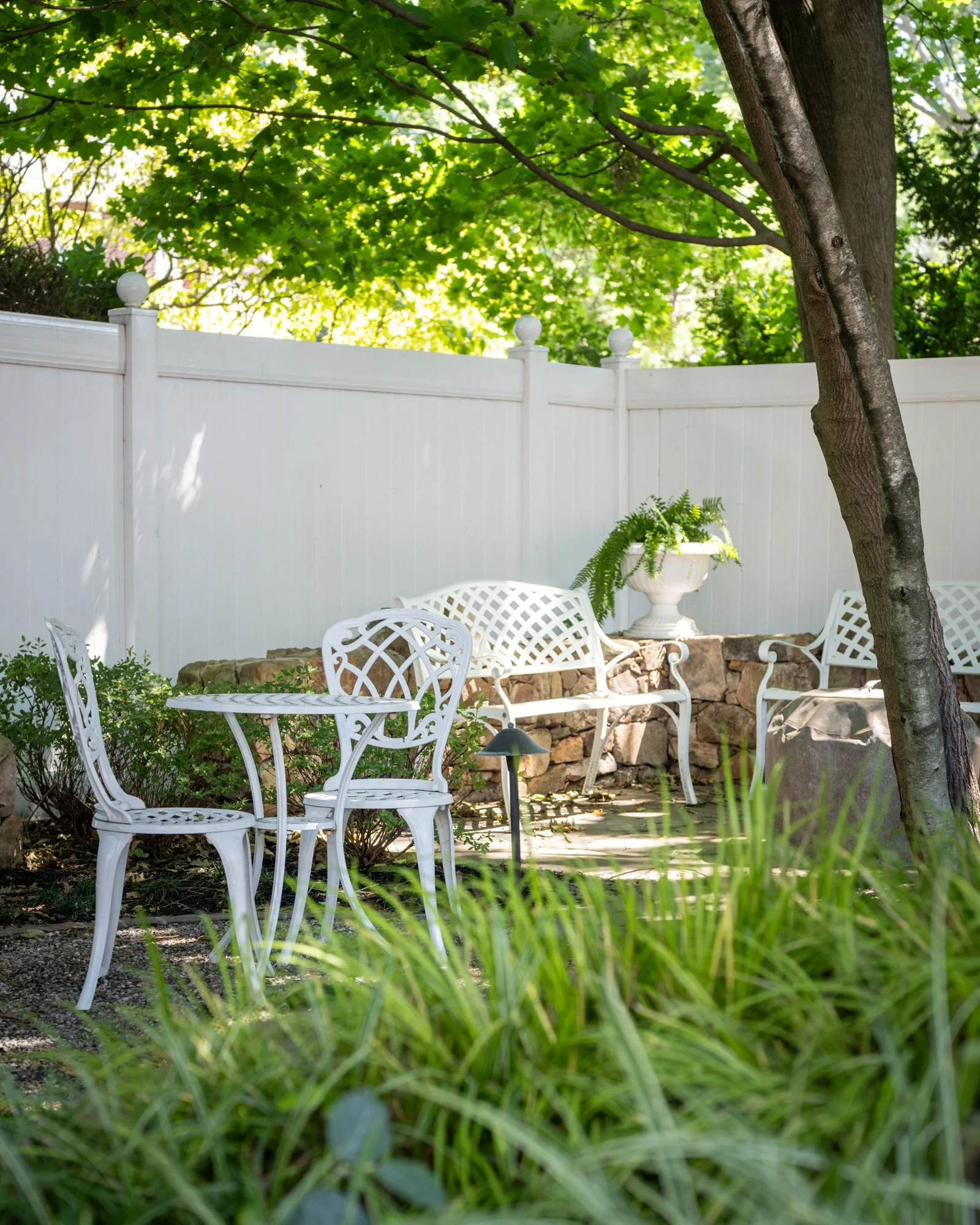 A backyard patio area with white metal outdoor furniture including chairs and a table, surrounded by green plants, trees, and a white privacy fence, with sunlight filtering through the leaves.