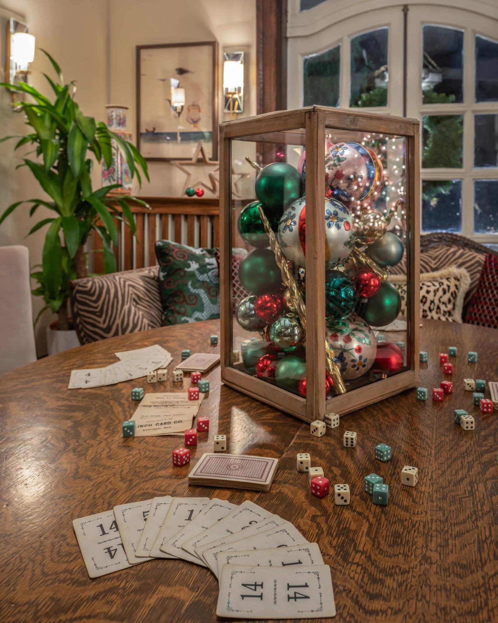 A tabletop playing card game setup with scattered playing cards and dice, a glass display case filled with multicolored Christmas ornaments, and a cozy room with a large plant, zebra print pillows, and artwork on the wall.