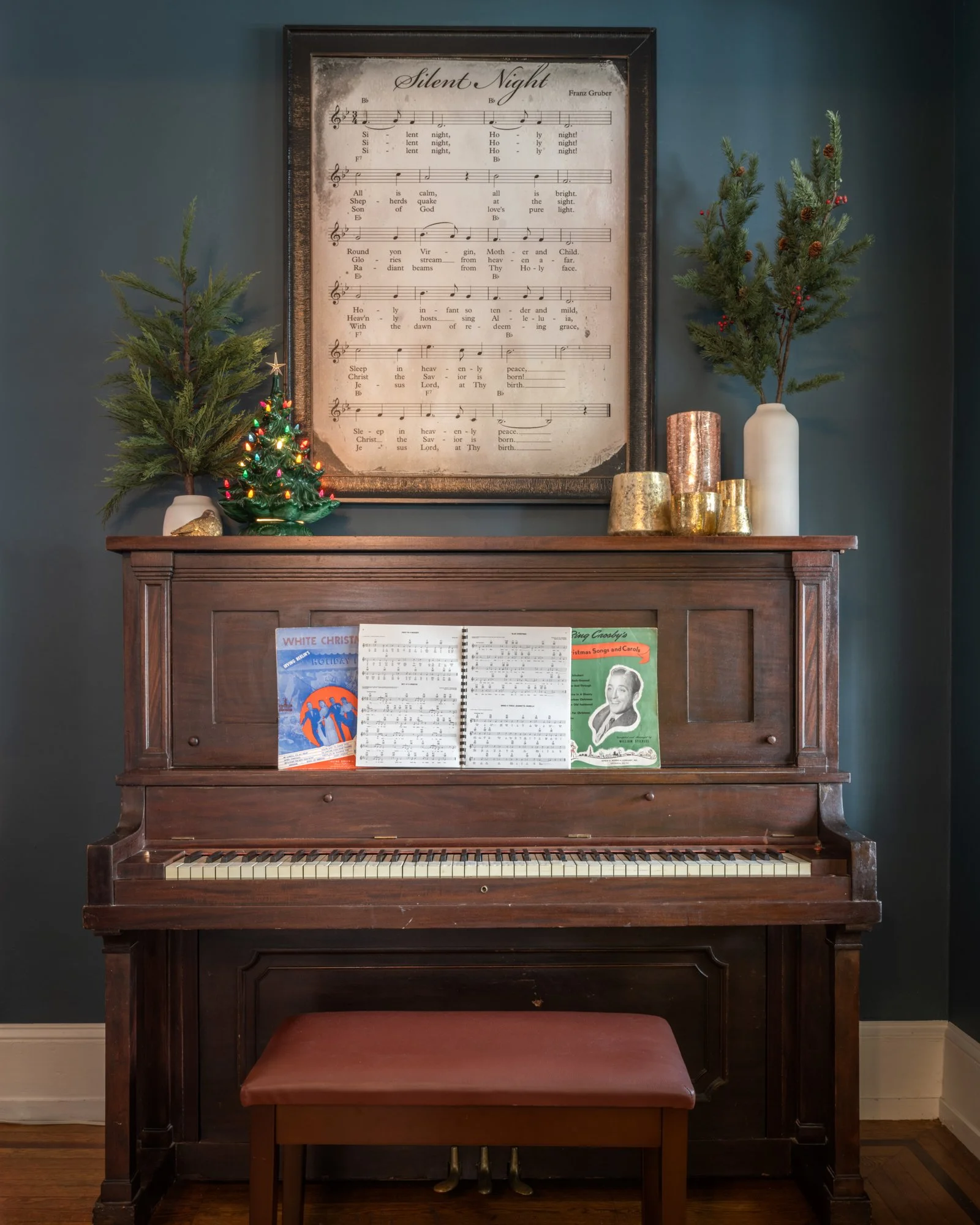 A dark wooden upright piano with sheet music on top, two small Christmas trees in white vases, and decorative candles on the piano. A framed sheet music titled 'Silent Night' hangs above the piano. Red berries adorn the right Christmas tree, and a sm