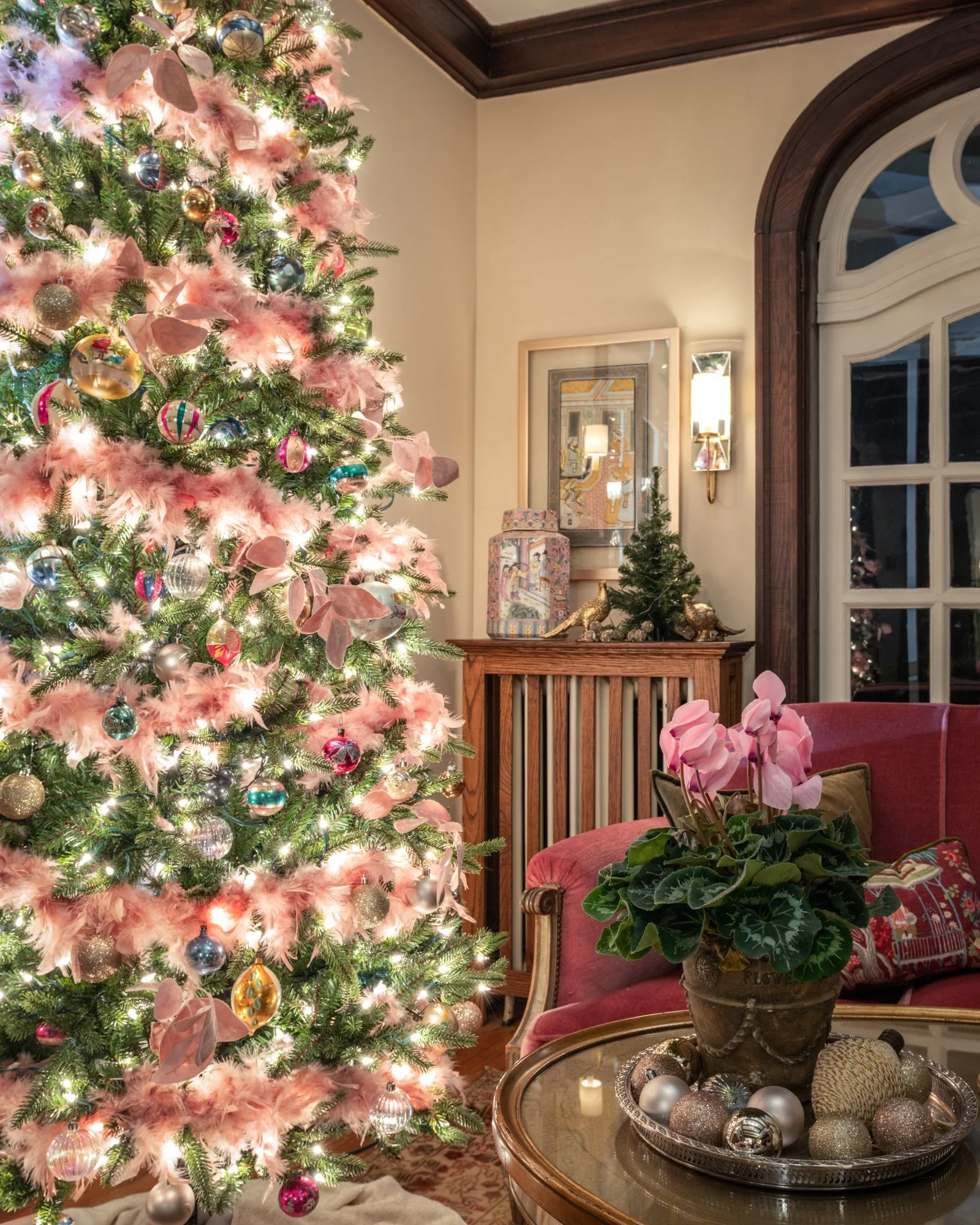 A decorated Christmas tree with pink feathers and multicolored ornaments in a living room, with a small decorated tree, framed artwork, a table with a silver tray holding Christmas ornaments and a potted plant, and a pink armchair.