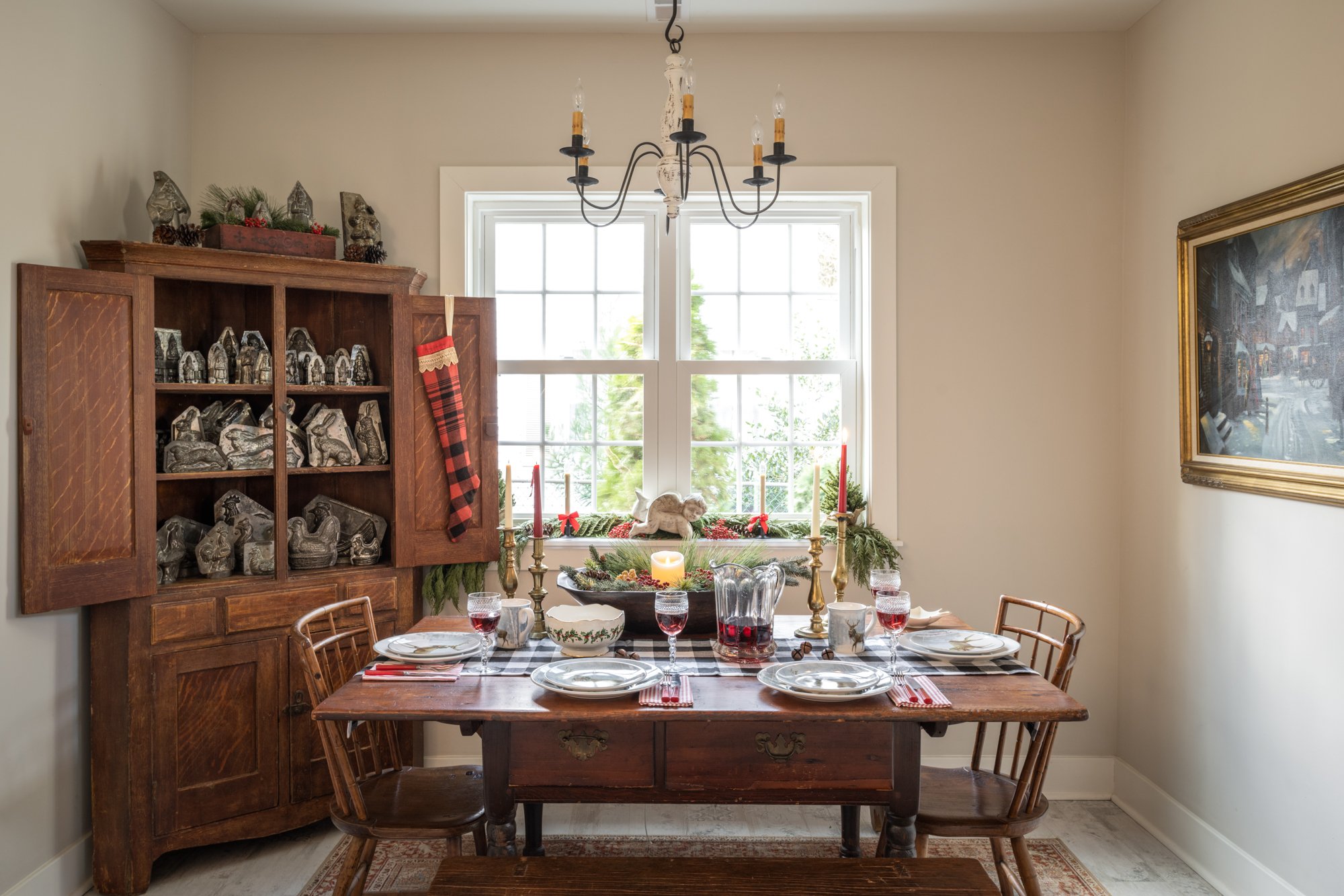 A dining room with a wooden table set for a Christmas meal, featuring plates, glasses, and napkins. The table has a decorative centerpiece with candles, pinecones, and greenery. There are four wooden chairs around the table. In the background, a wind