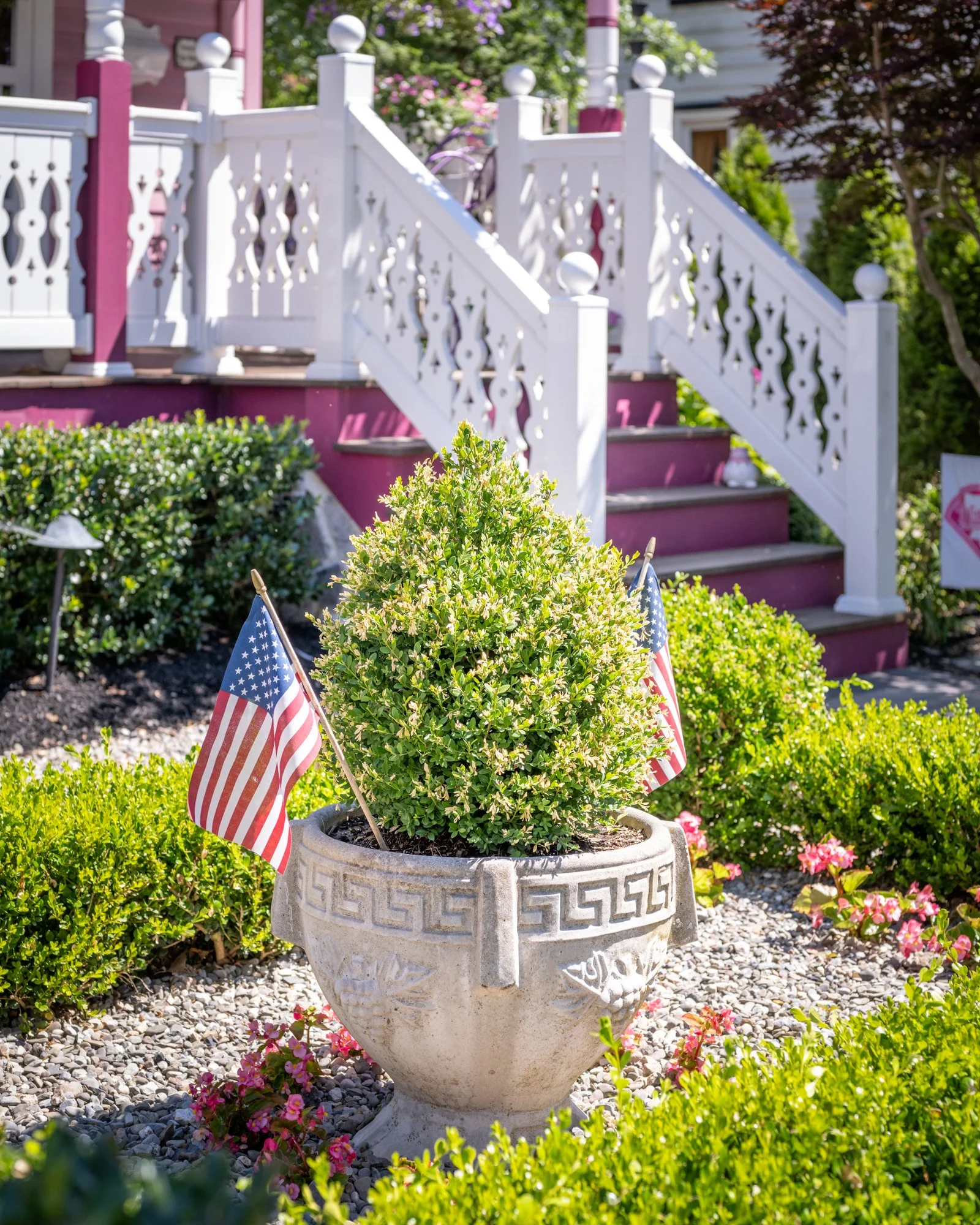 A white stone planter with a Greek key pattern around the top, holding a round, lush green bush with two small American flags on sticks. The planter is situated in a garden with pink and green flowering plants and trimmed bushes, with a backdrop of a
