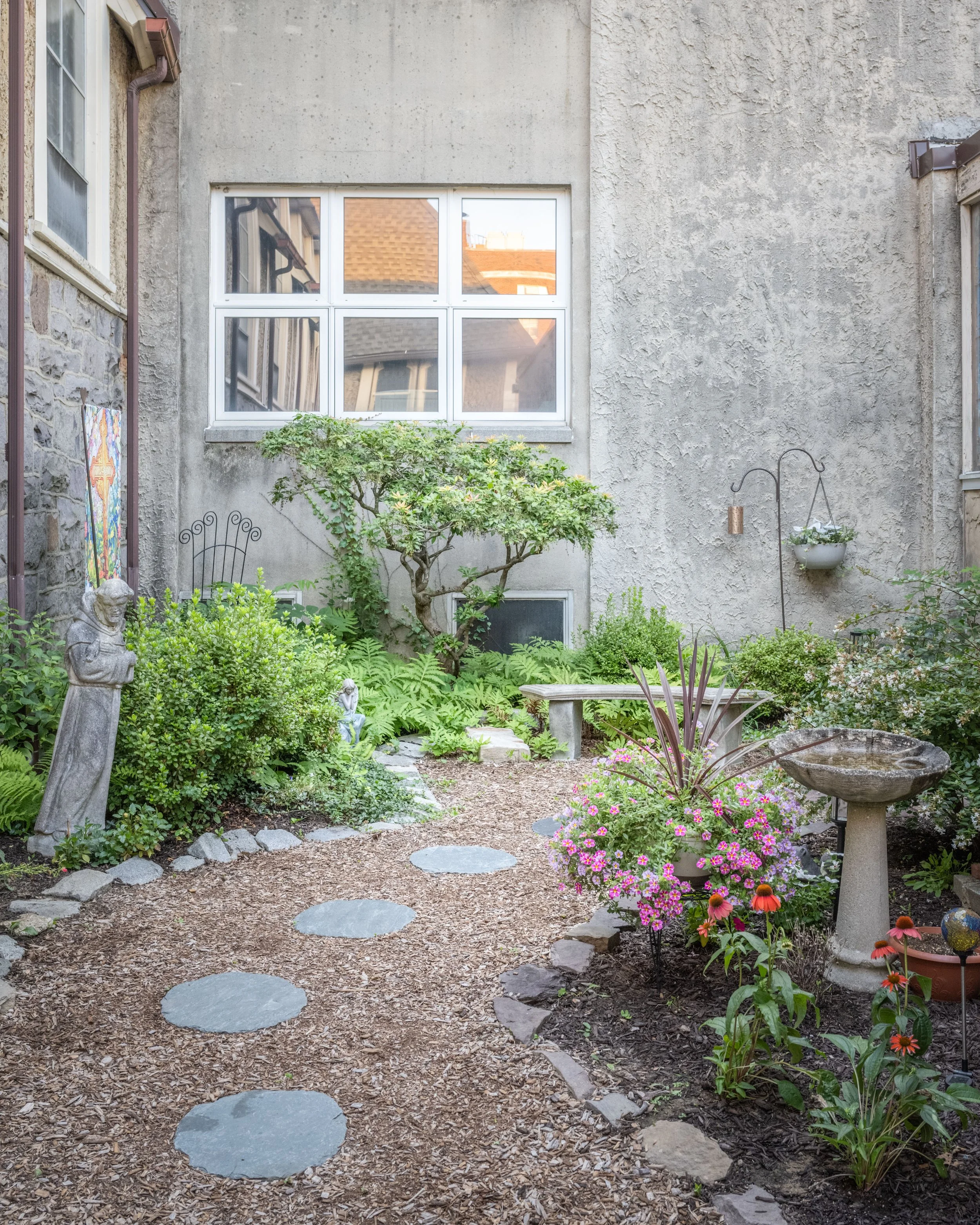 A backyard garden with a stepping stone path, various green plants, blooming flowers, garden statues, and a stone birdbath, with a house's exterior wall with a window in the background.