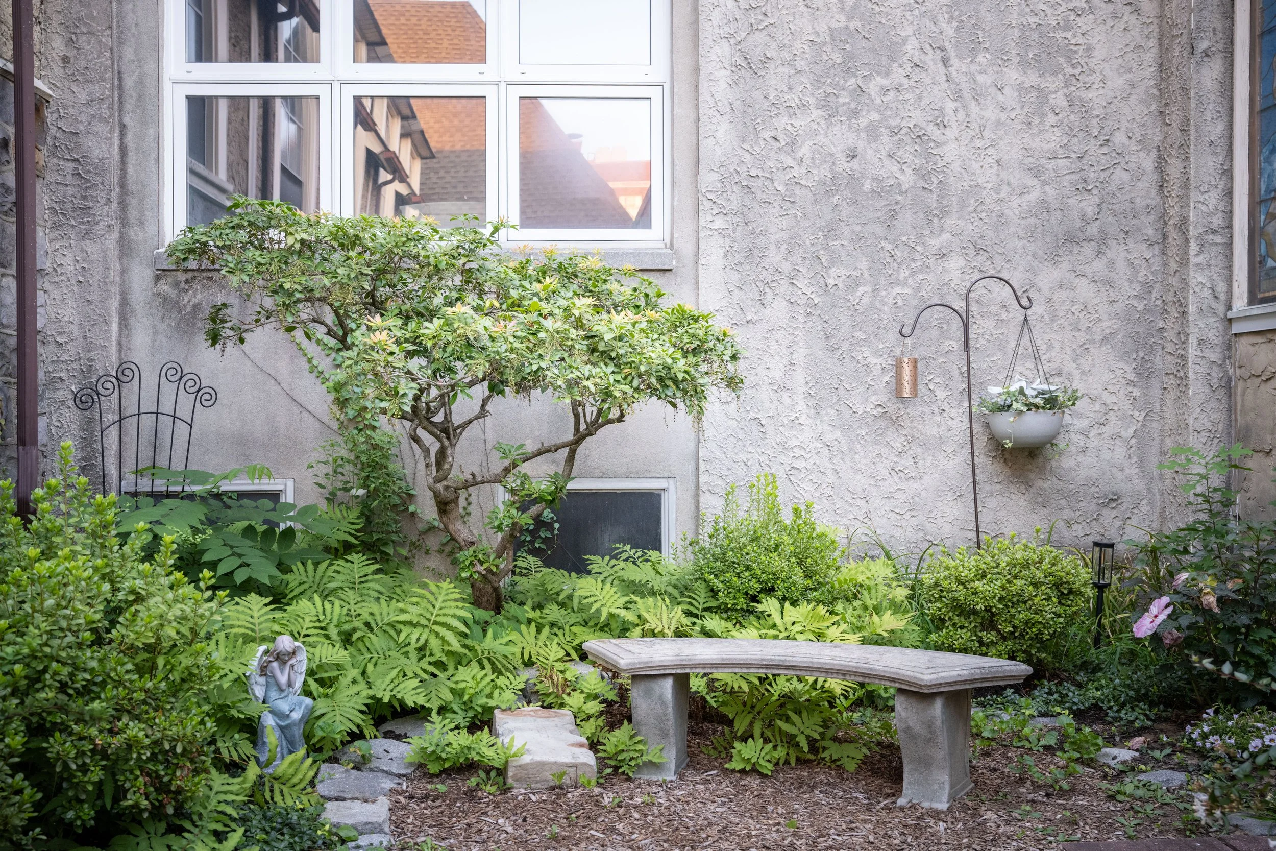 A small garden with lush green plants, a tree, a statue of an angel, and stone benches, with a house wall and window in the background.