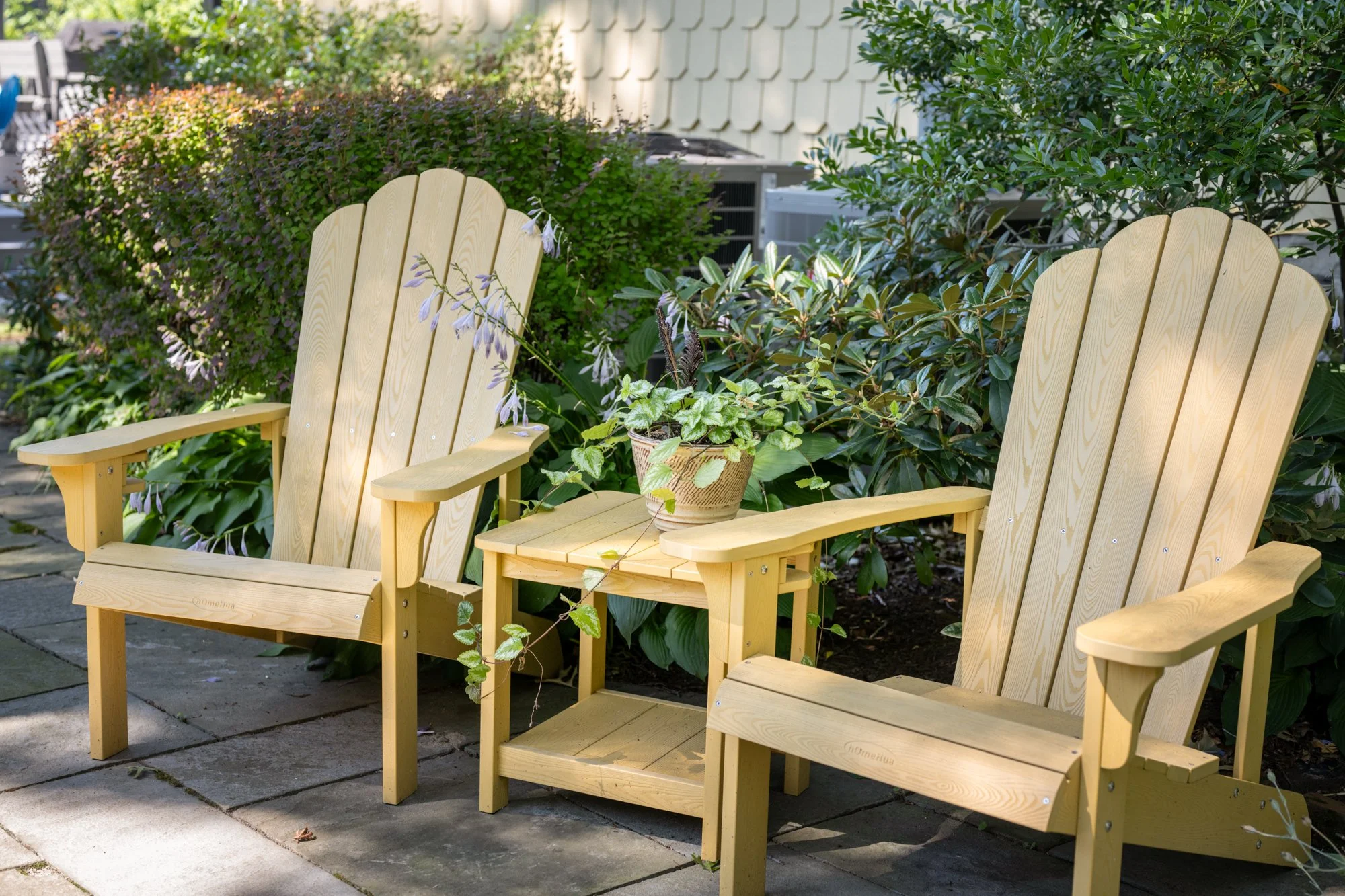 Two wooden Adirondack chairs in a garden with green bushes and potted plants, on a patio with stone paving.