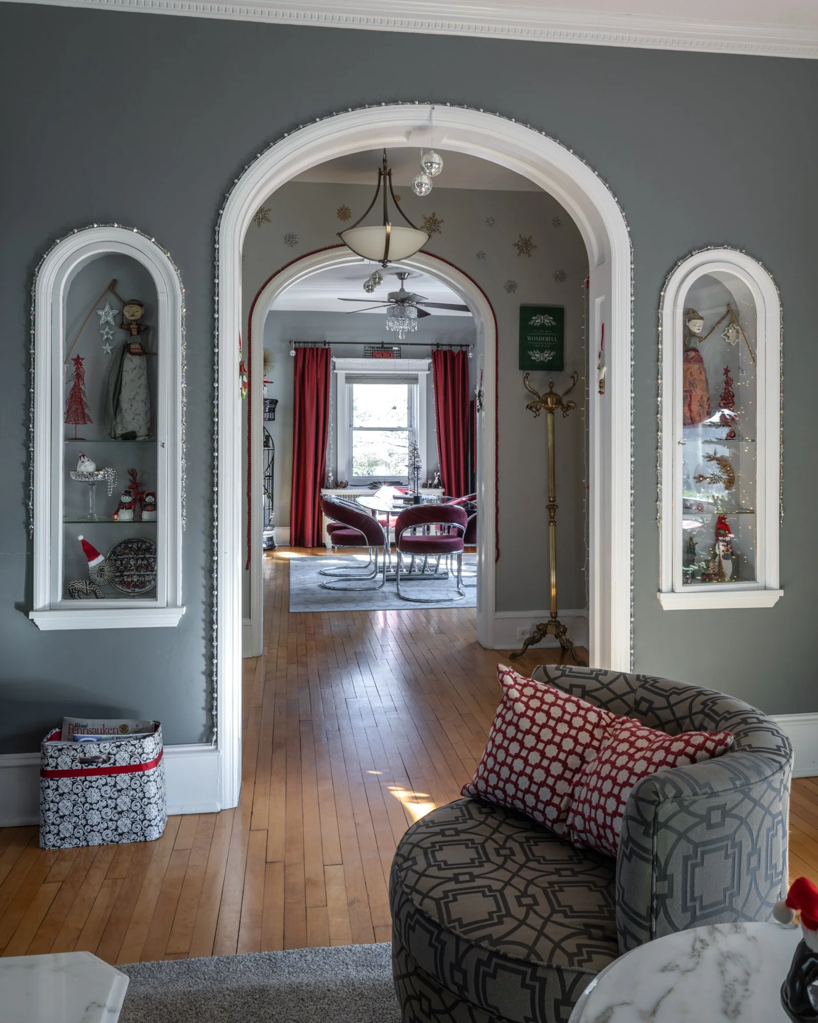 Living room with a view into a dining area, decorated for Christmas with red curtains, snowflake ornaments, and holiday decorations. There's a patterned sofa with red and gray pillows, a marble-topped side table, and built-in wall display cabinets wi