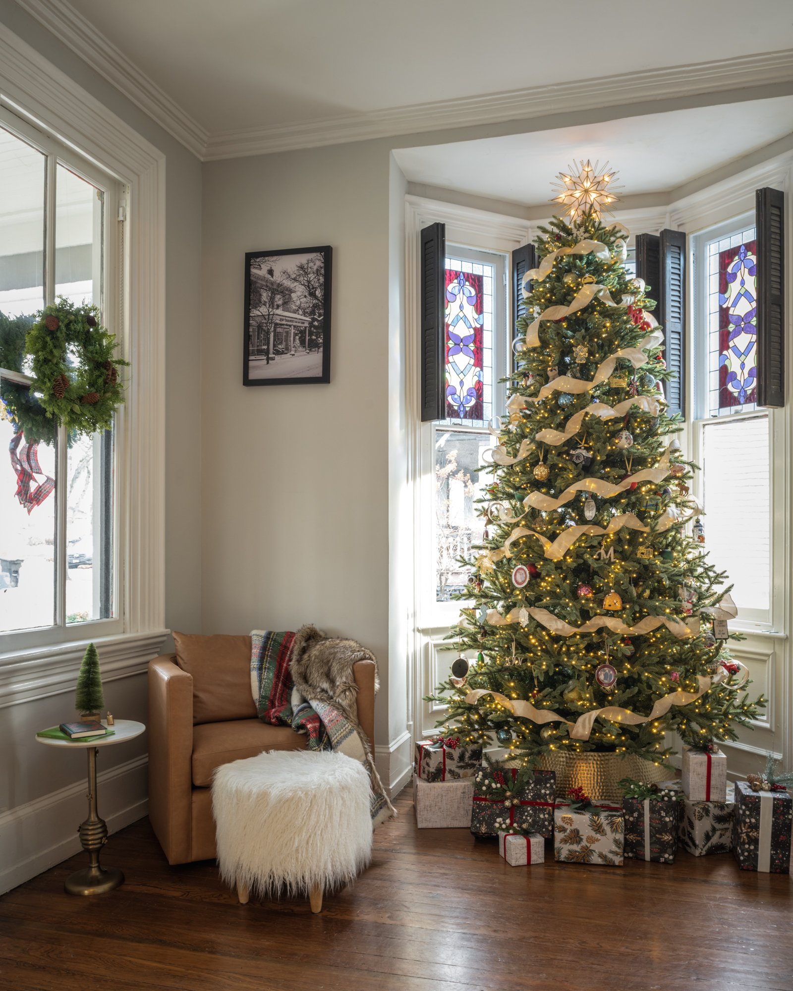 A decorated Christmas tree with lights, ornaments, and a star topper in a living room. Presents are placed under the tree. Nearby, a tan armchair with a blanket and a furry cushion, a small side table with a book and a small plant, and windows with C