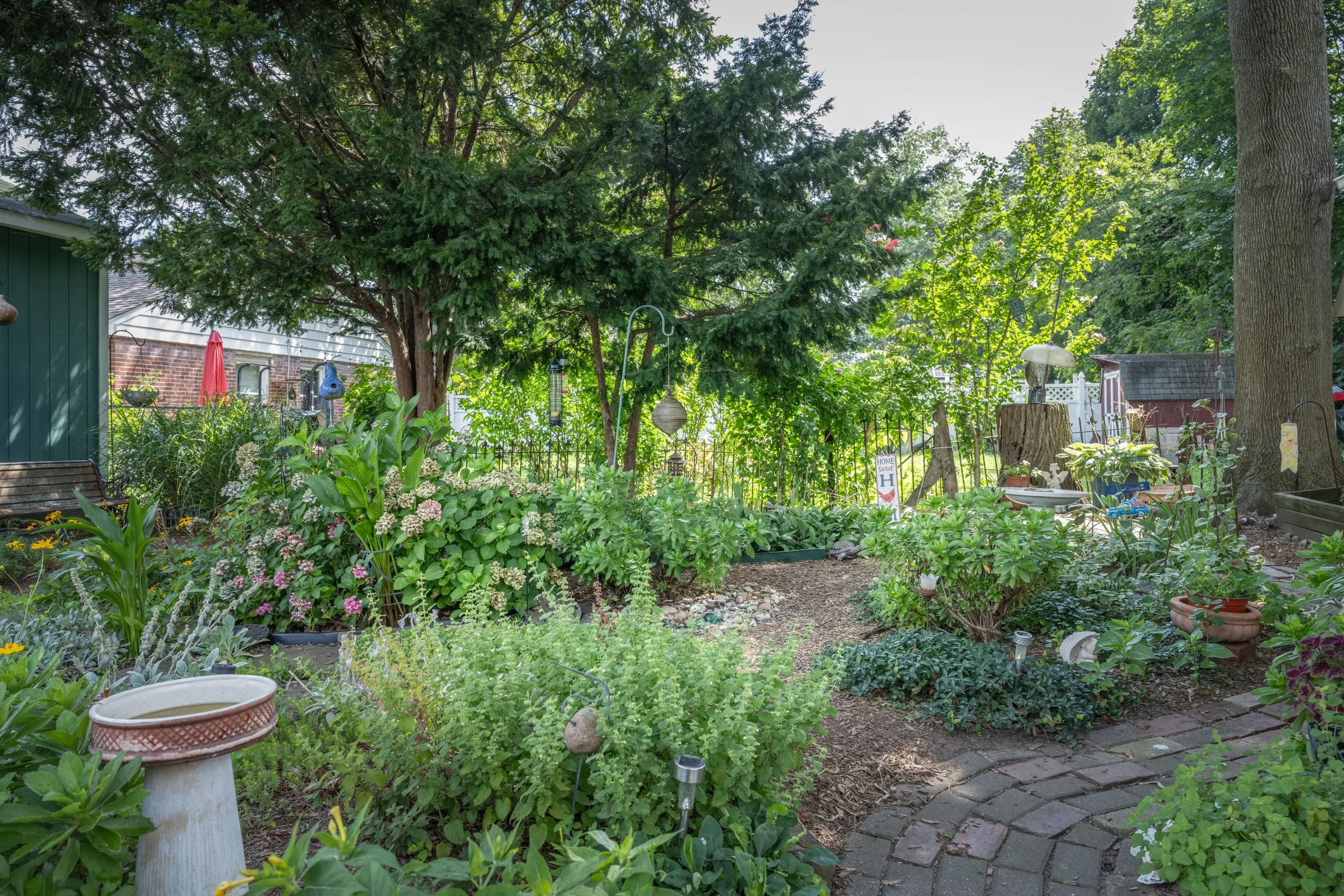 A lush green backyard garden with various plants and flowers, surrounded by trees and garden decorations, and a brick house in the background.